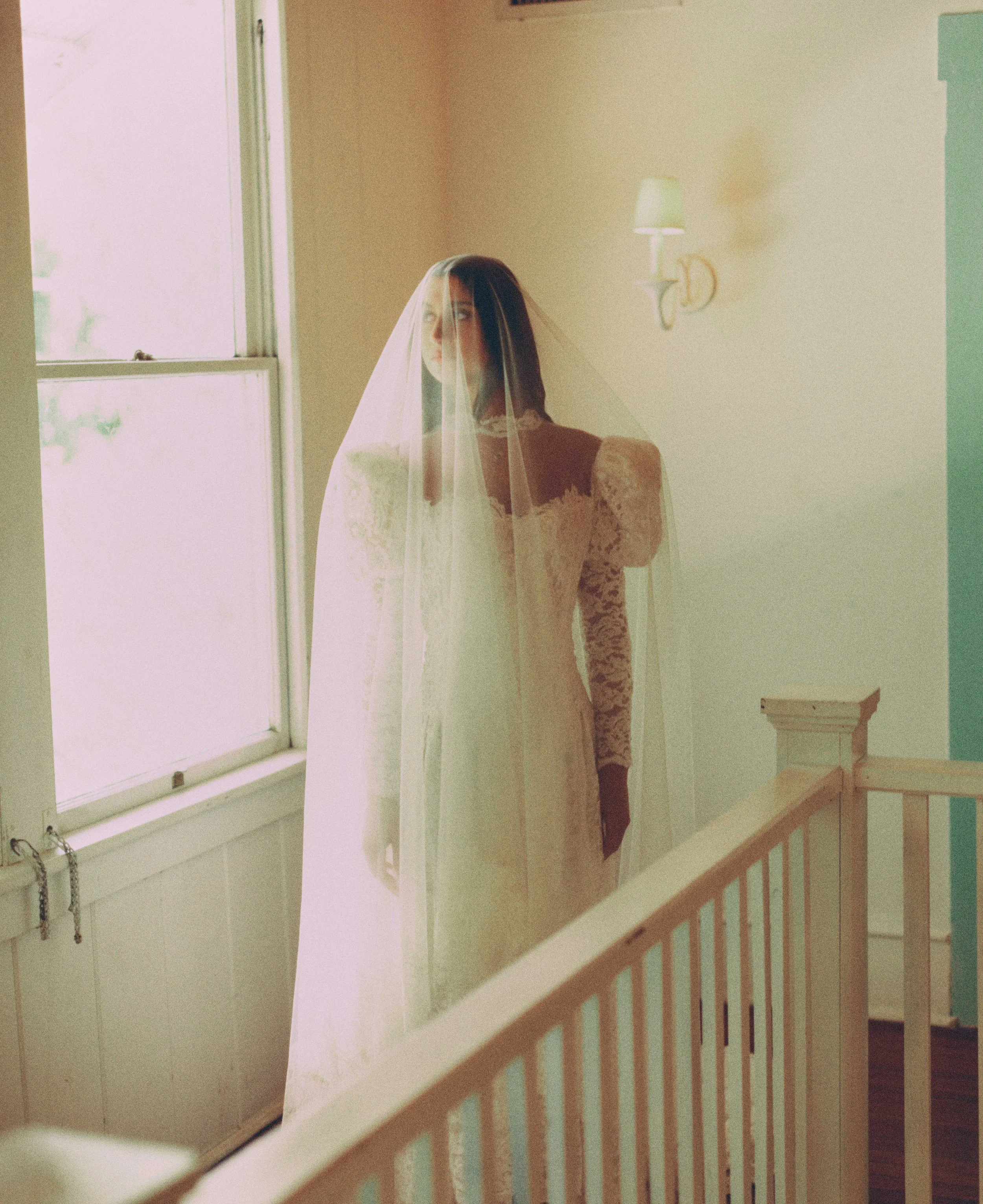 A woman with dark hair wearing a wedding dress and veil, standing behind a glass window with foliage outside, inside a room with cream walls and a small wall lamp.