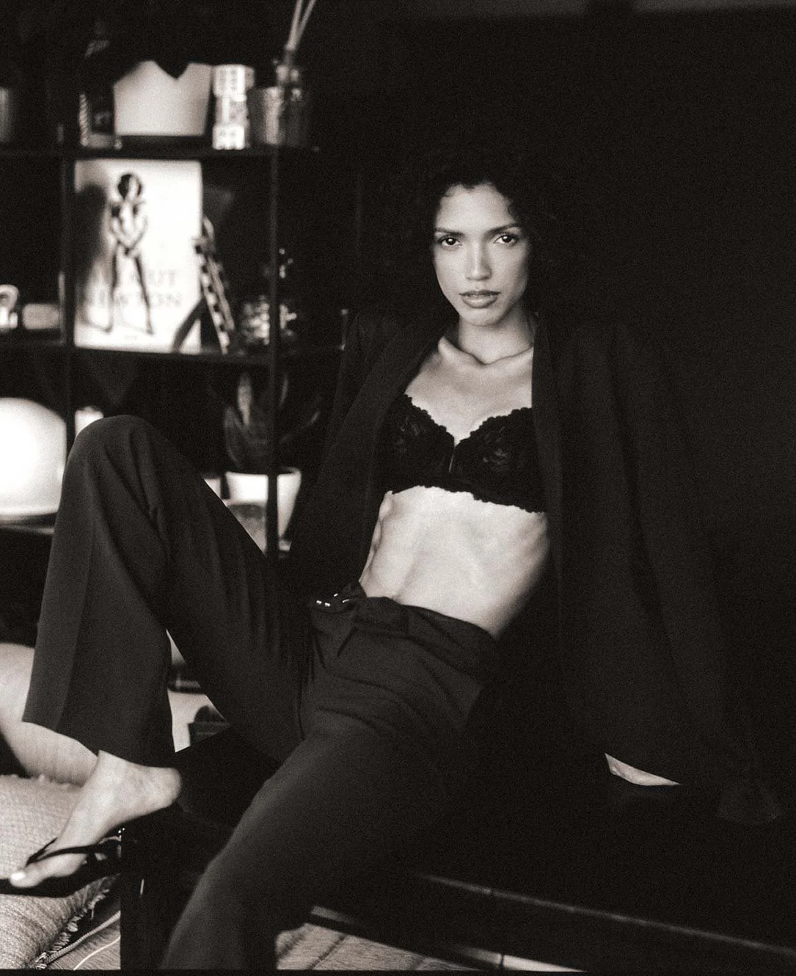 A black and white photograph of a woman sitting on a bench in a room. She is wearing a black lace bra, black pants, and a dark blazer. Her hair is curly and she is looking directly at the camera with a serious expression. In the background, there is 