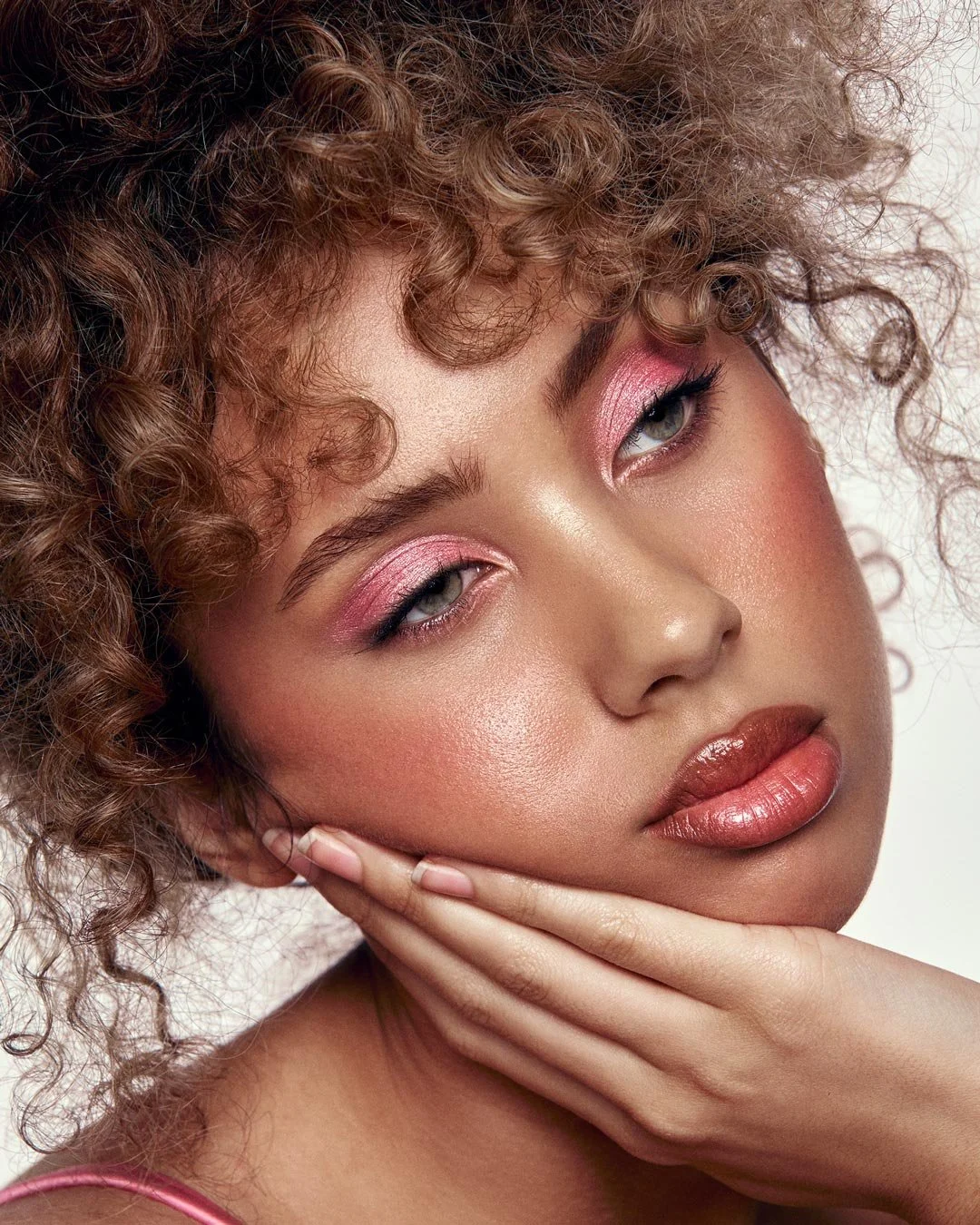 Close-up of a woman with curly hair, pink eyeshadow, glossy lips, and a neutral expression, resting her chin on her hand.