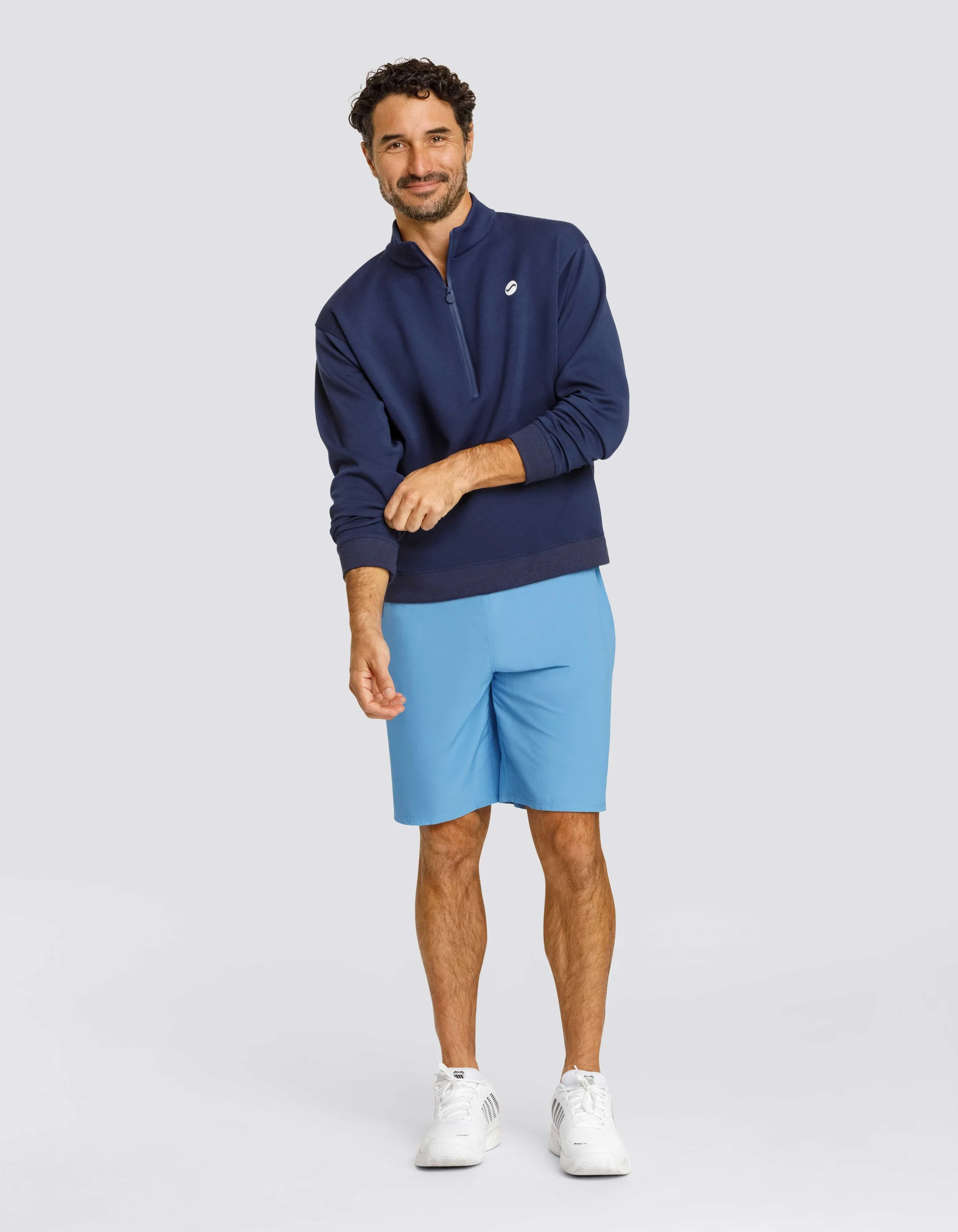 A man with curly dark hair and a beard posing against a light gray background, wearing a navy blue quarter-zip athletic top, light blue shorts, and white sneakers.