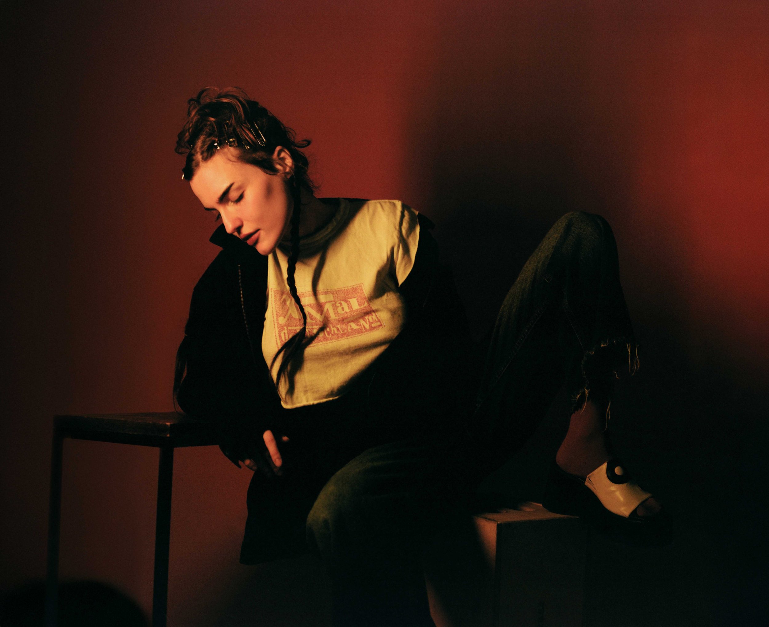A woman with curly hair styled with hairpins, wearing a yellow t-shirt and dark jacket, sitting on a black chair against a dark red background. She has her eyes closed and appears to be in a relaxed or contemplative mood.