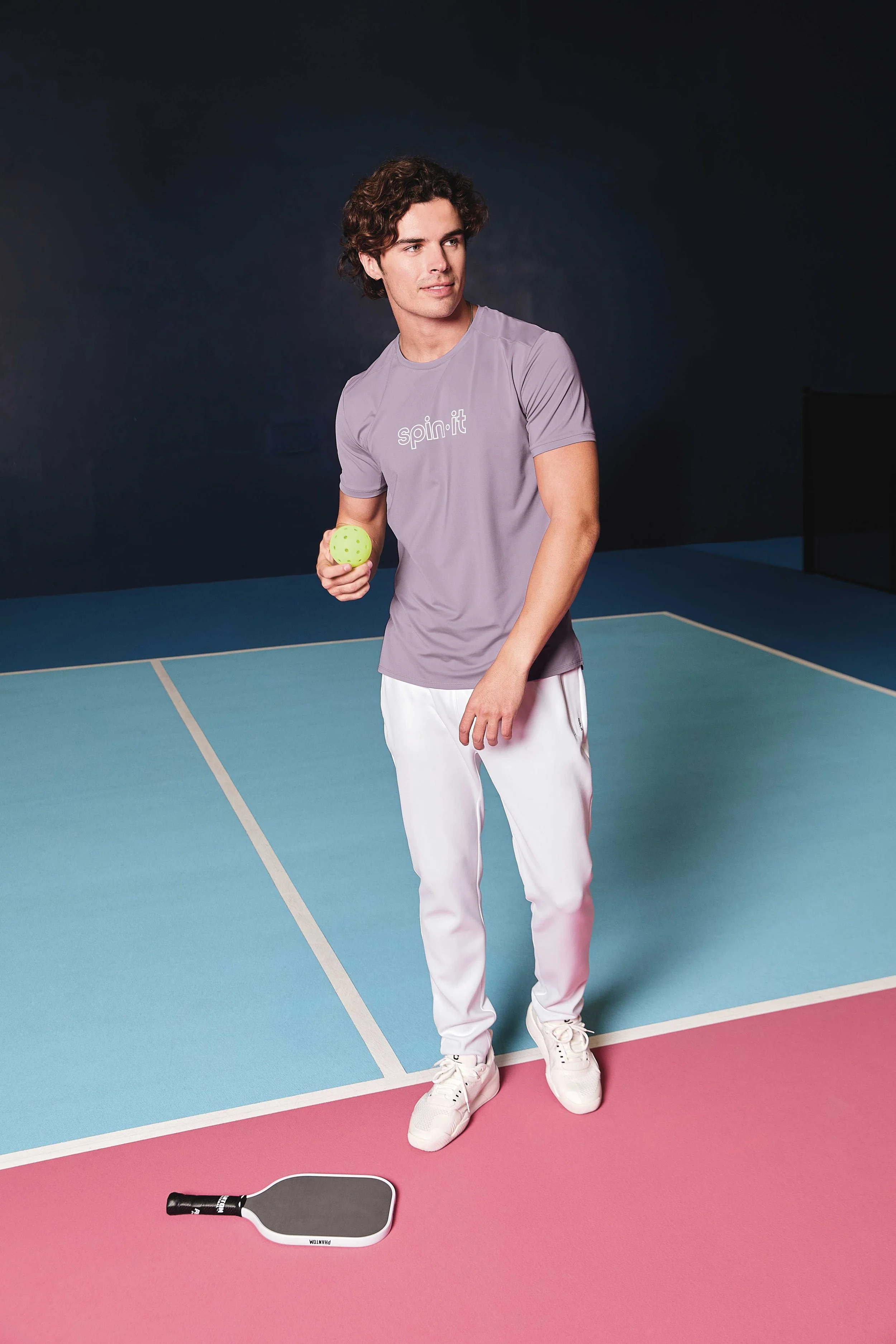 A young man with curly hair wearing a purple 'spin it' T-shirt and white pants stands on a tennis or pickleball court, holding a green pickleball paddle. The court has light blue and pink sections, and there is a black paddle lying on the pink surfac