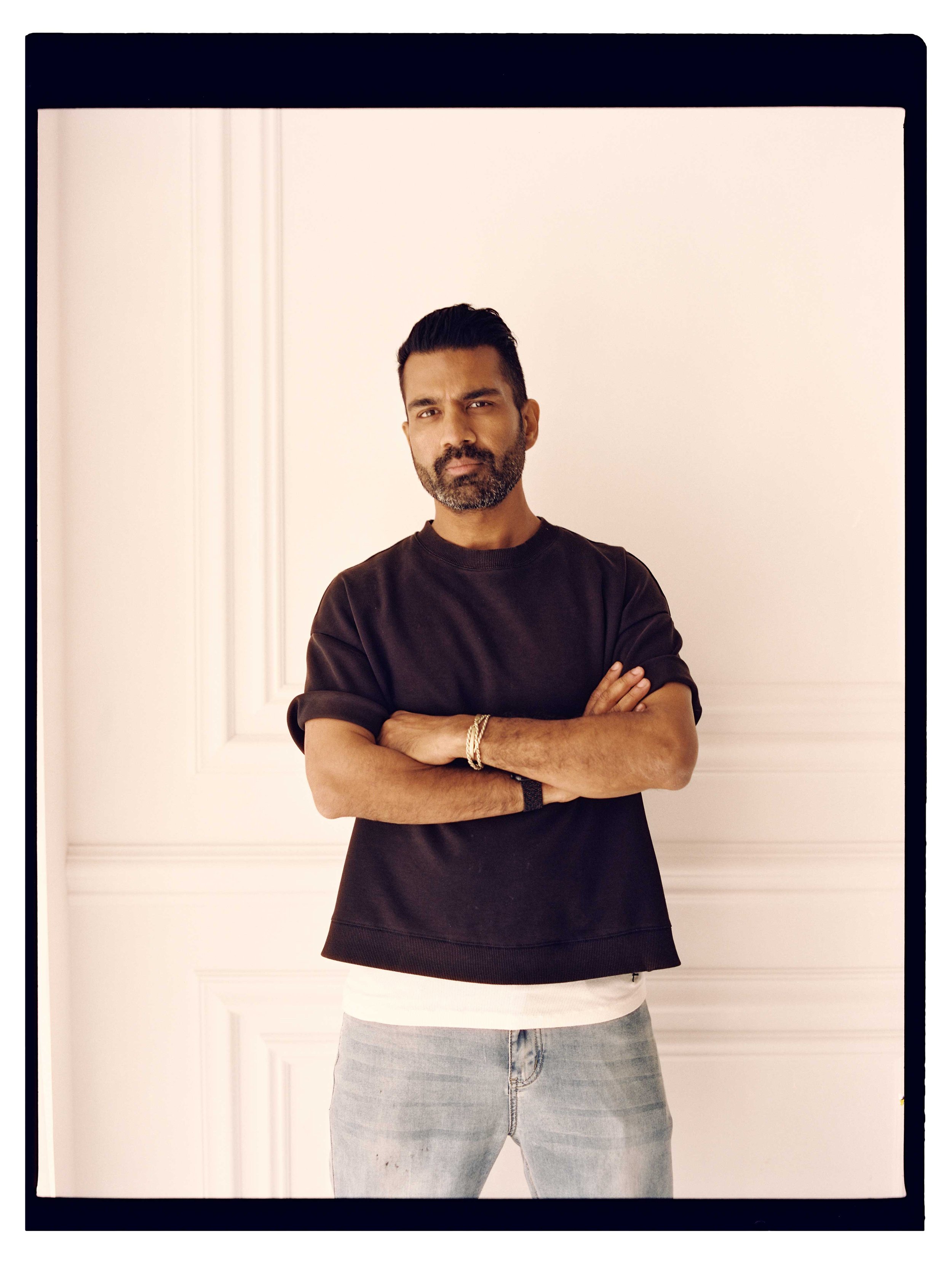 A man with dark hair, beard, and tan skin wearing a black sweatshirt with arms crossed standing in front of a plain white wall.