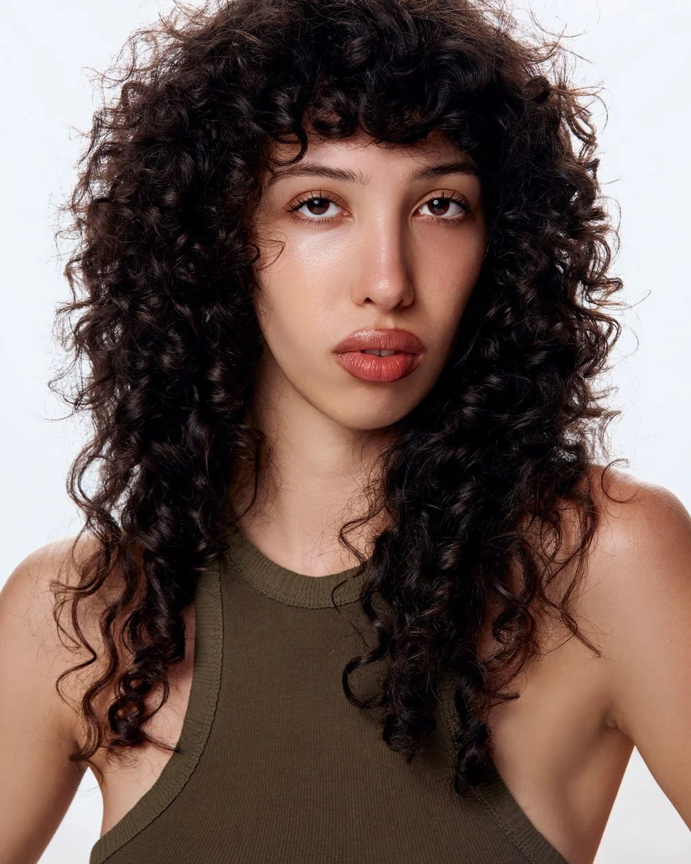 A woman with curly dark hair and light skin, wearing a sleeveless olive green top, looking directly at the camera against a plain white background.