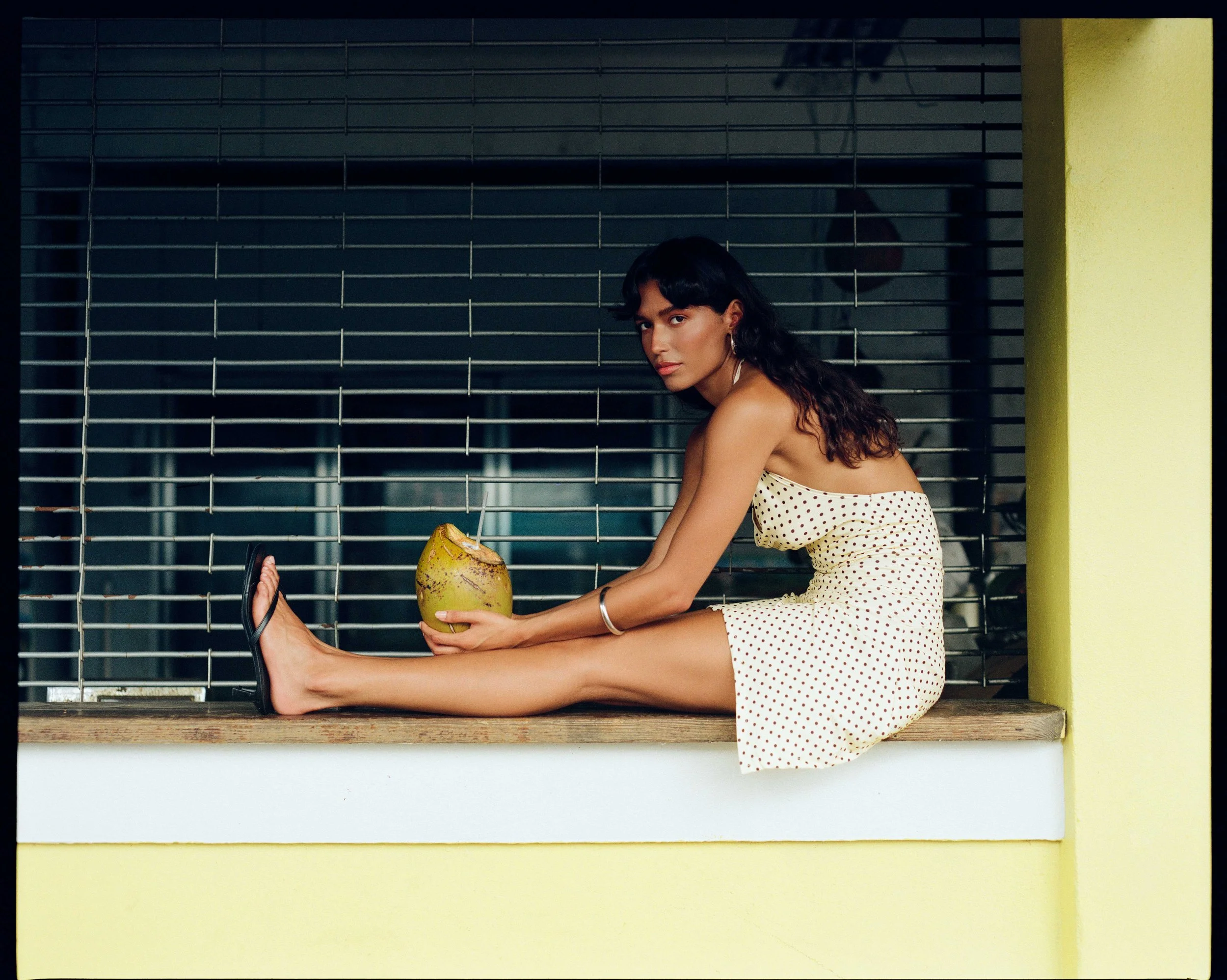 A woman in a polka dot dress sitting on a windowsill holding a green coconut with a straw, looking at the camera with a serious expression. The window has metal bars and a yellow wall surrounding it.