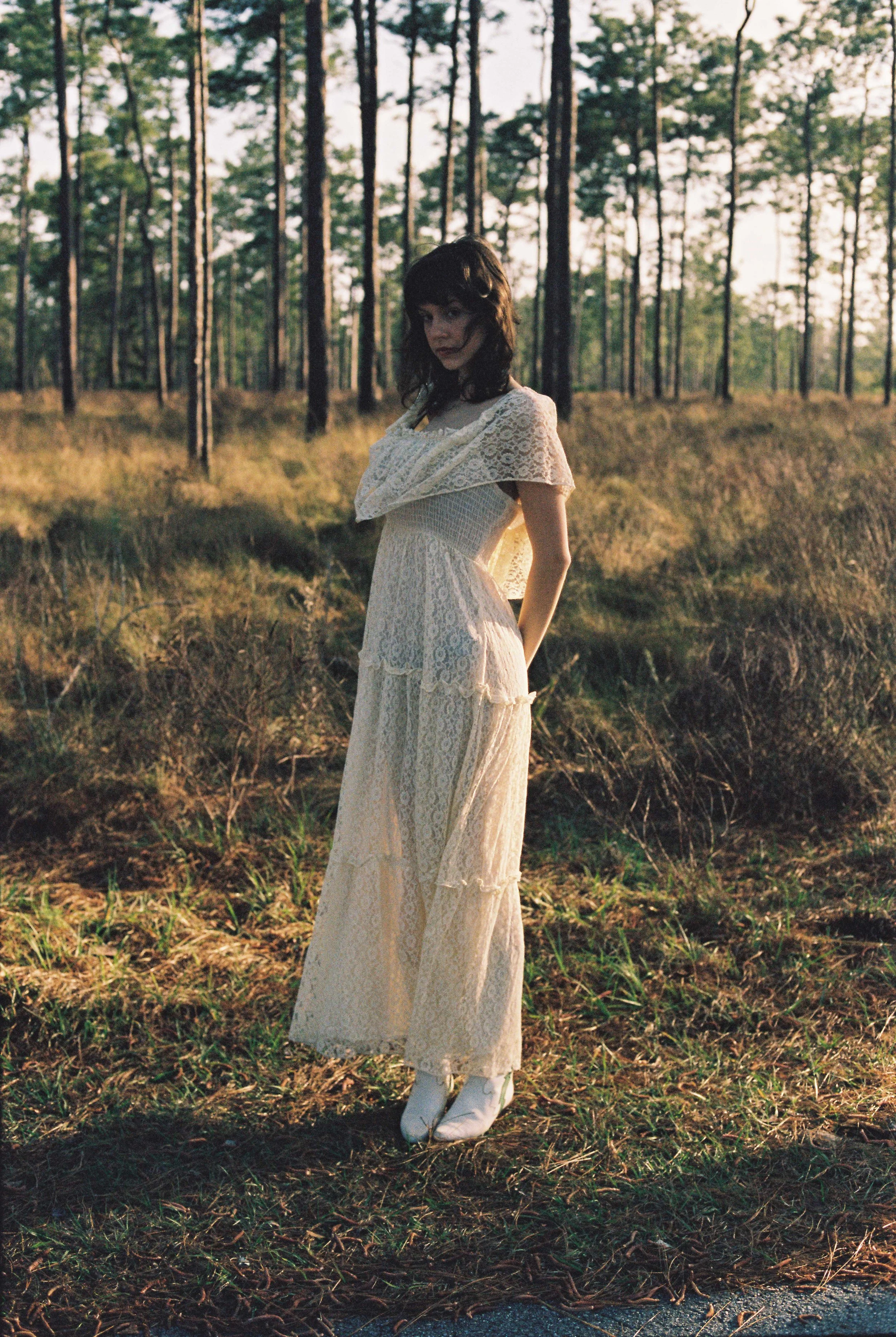 A young woman with dark hair wearing a white lace dress, standing in a forest during sunset.