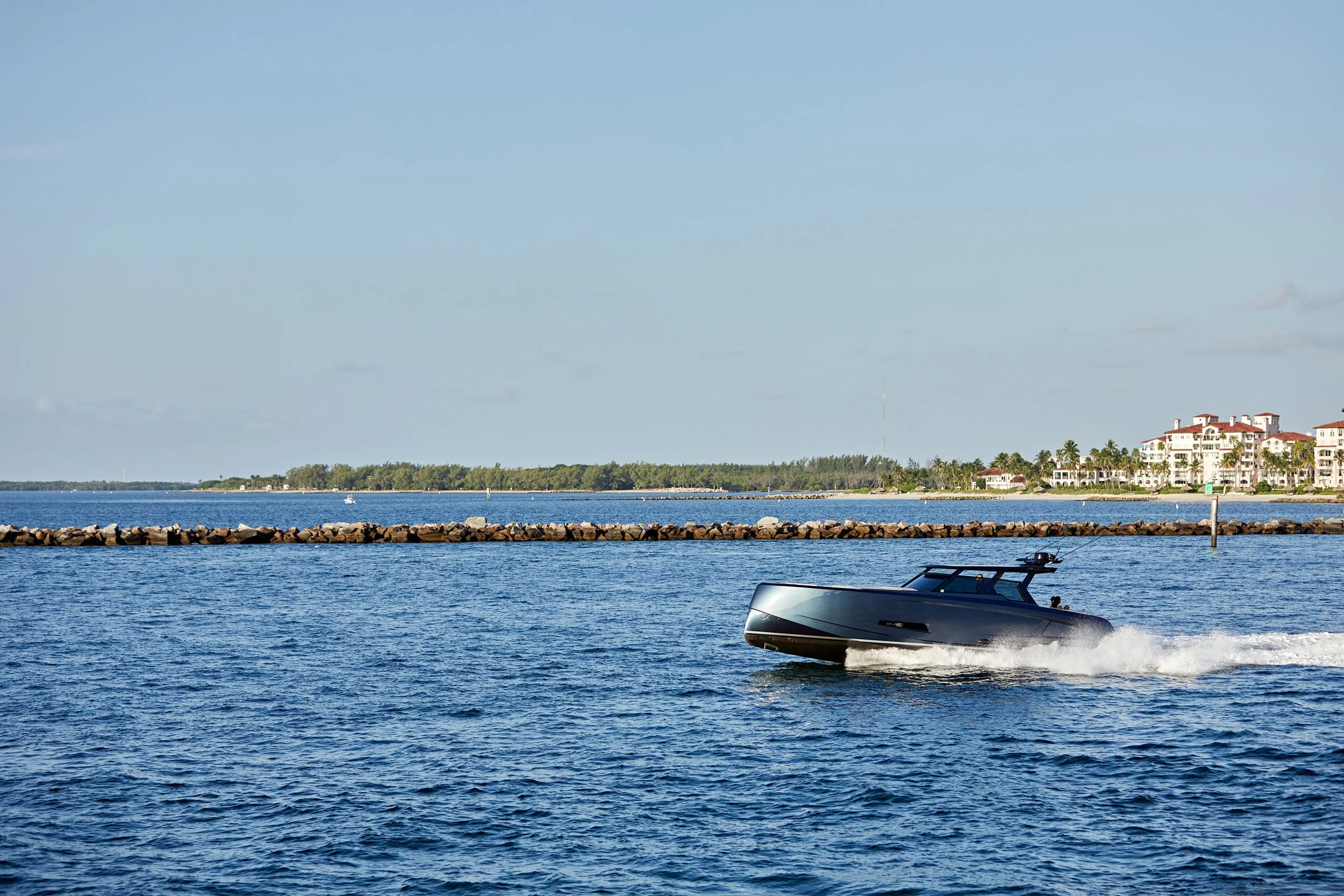 A boat cruising on calm waters near a breakwater with buildings and trees in the background under a clear blue sky.