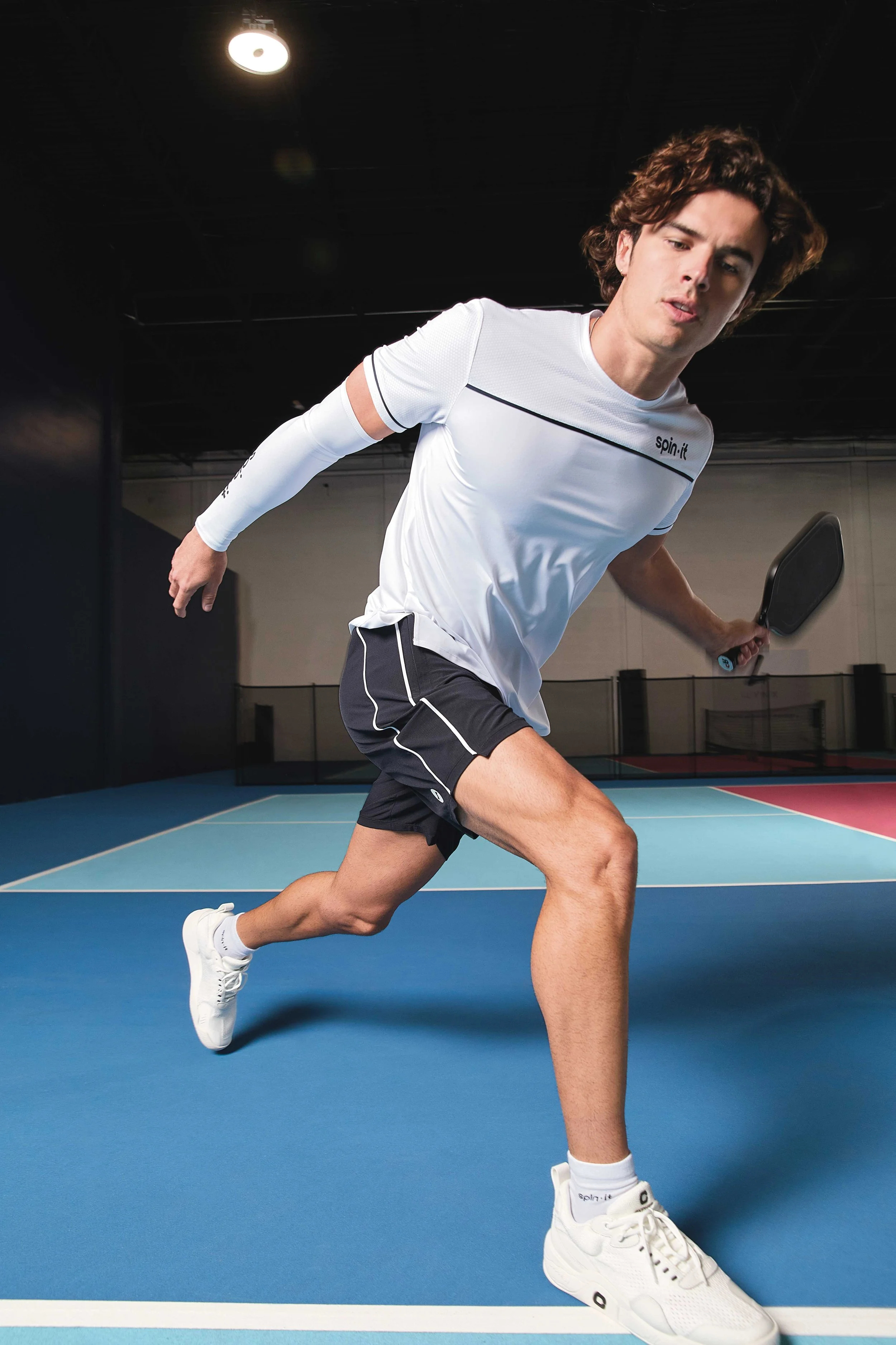 A young man playing pickleball on a blue indoor court, wearing a white athletic shirt, black shorts, white sneakers, and holding a pickleball paddle, with a focused expression.