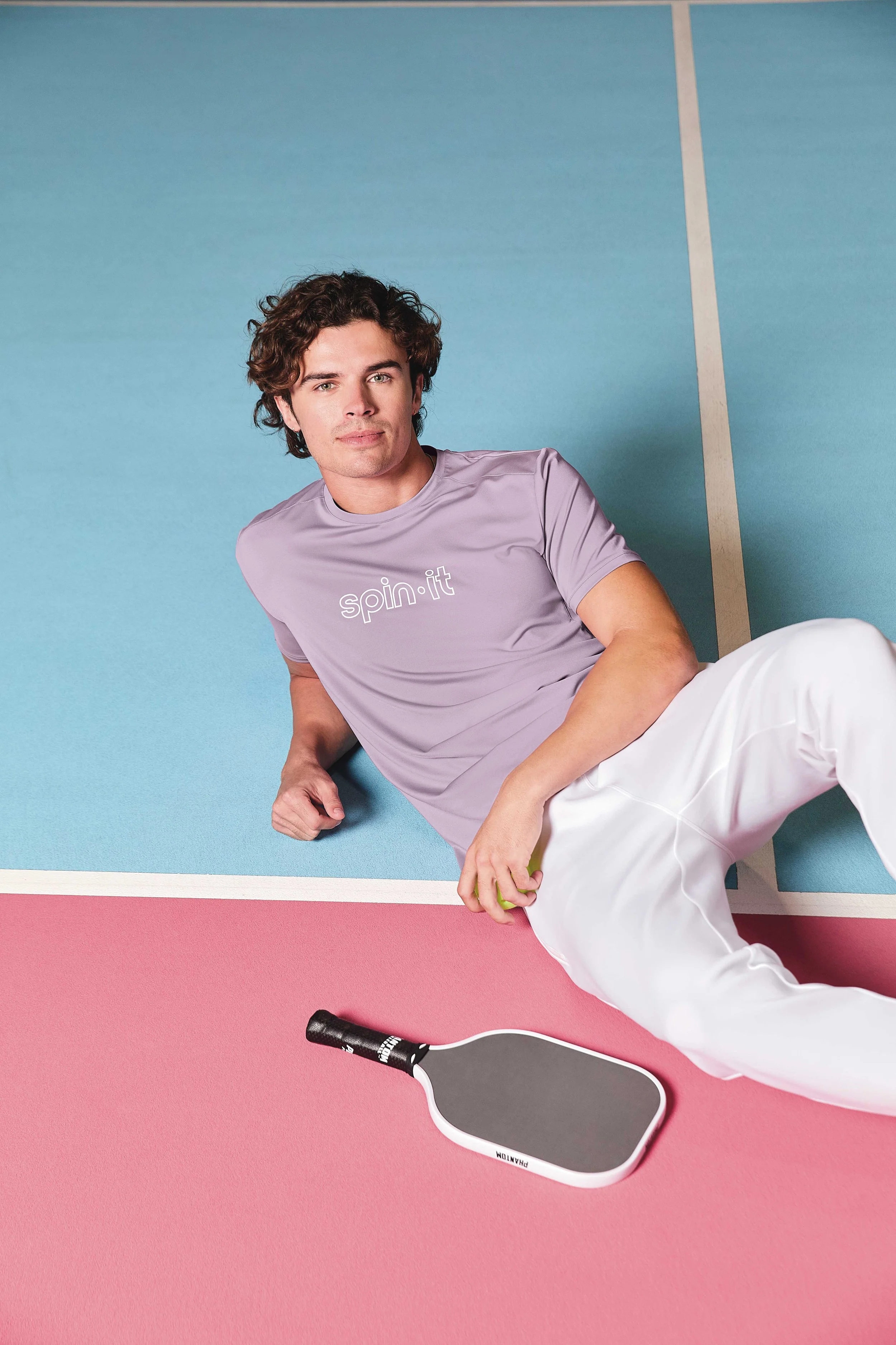 Young man with curly brown hair lying on a pastel tennis court with a pink and blue background, wearing a lavender t-shirt with the word "spin.it" and white pants, next to a black pickleball paddle.