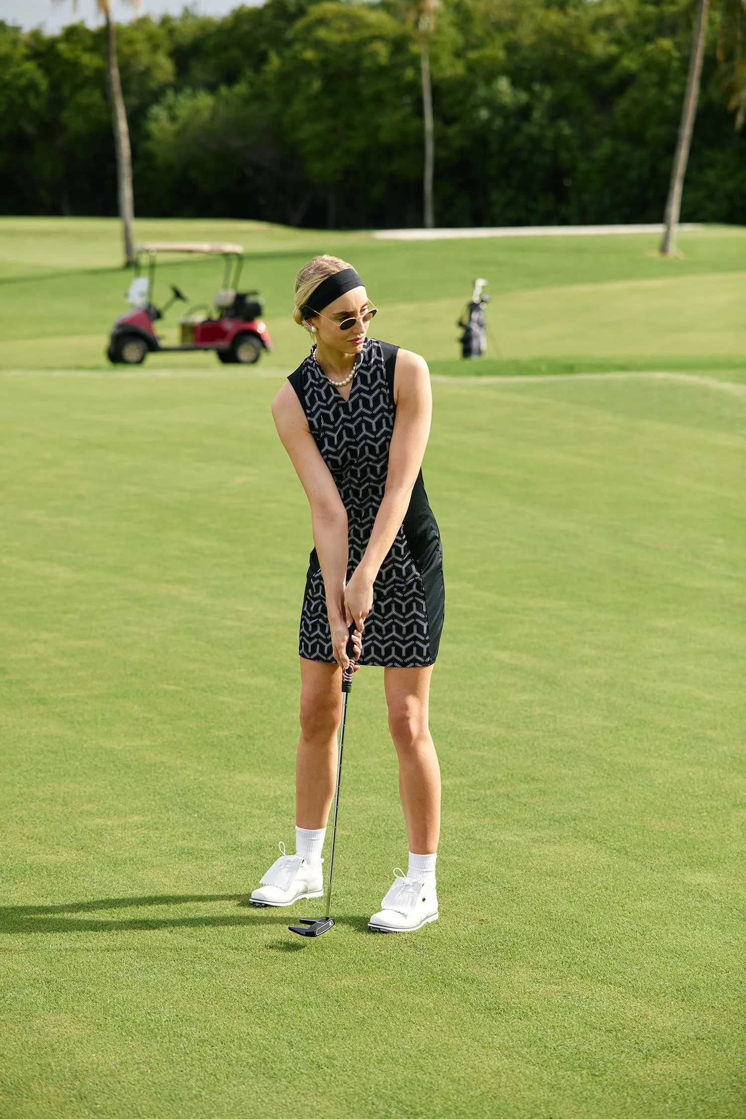 A woman dressed in vintage golf attire stands on a golf course holding a golf club, with a golf cart and trees in the background.