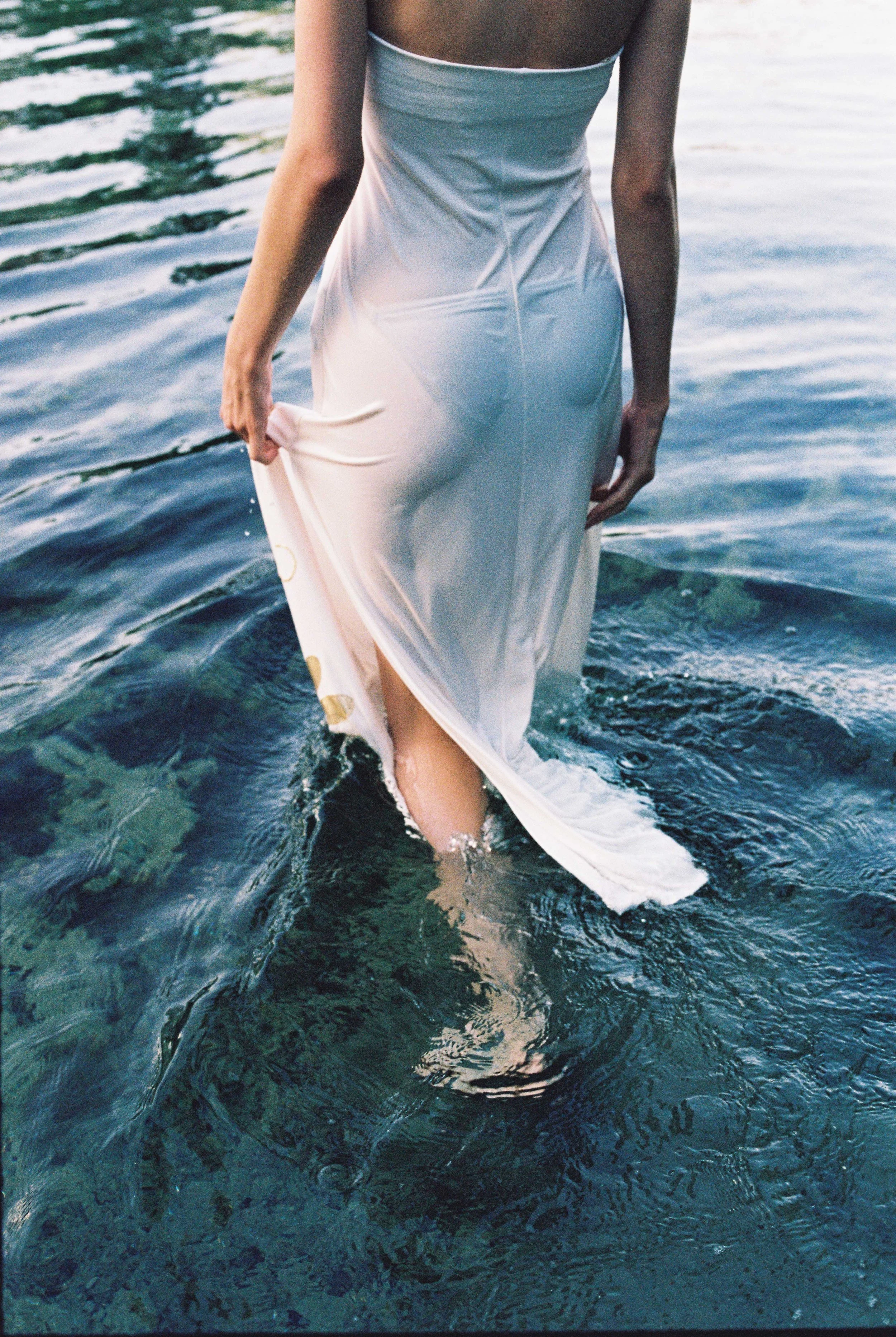 A person in a white dress walking in water, holding the dress with one hand and the lower part of their body submerged in the water.