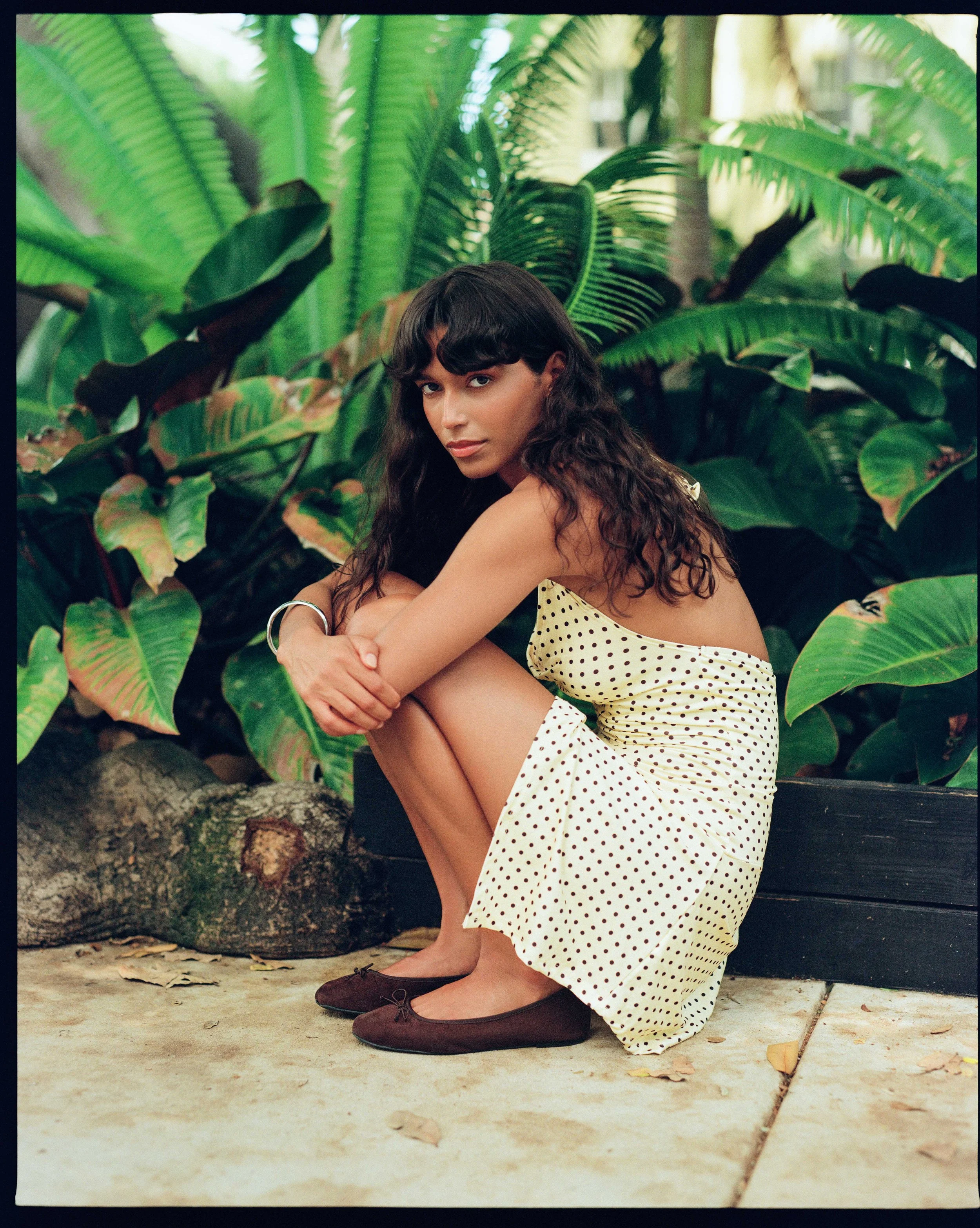 A woman with long, wavy dark hair sitting on a concrete ledge outdoors among large green tropical plants, wearing a cream-colored, polka-dotted dress and dark ballet flats, with her arms wrapped around her knees, looking at the camera.