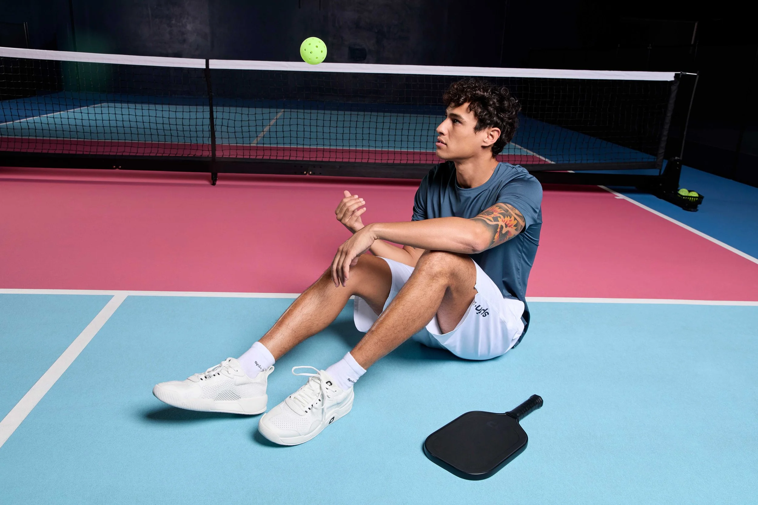 A young man sitting on the floor of an indoor pickleball court, resting with his knees bent, wearing a blue t-shirt, white shorts, and white sneakers. A pickleball is in the air above him, and a paddle is on the ground nearby. The court has pink and 