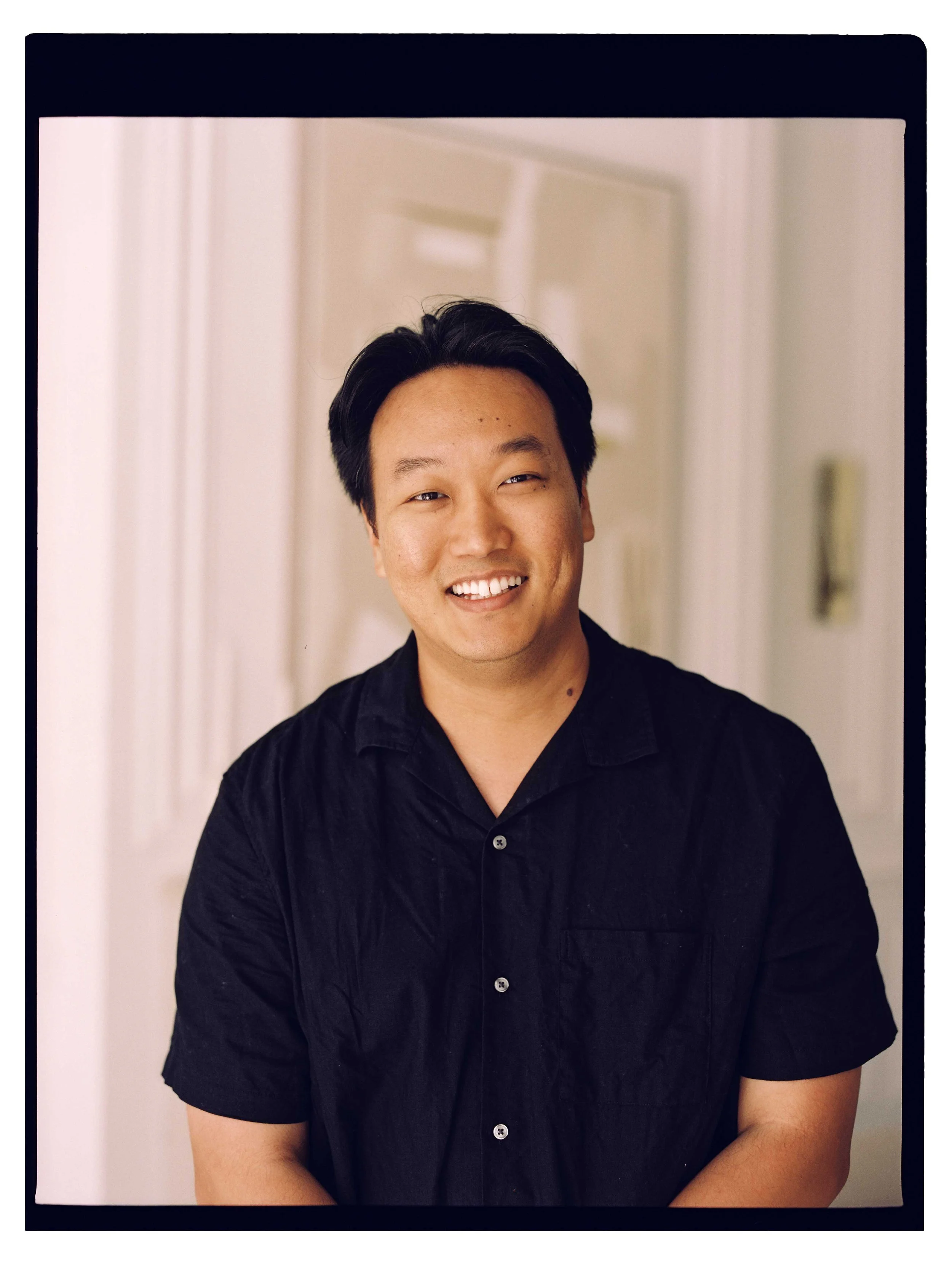 A young Asian man with short black hair smiling, wearing a black button-up shirt, standing in front of an off-white wall with a door in the background.