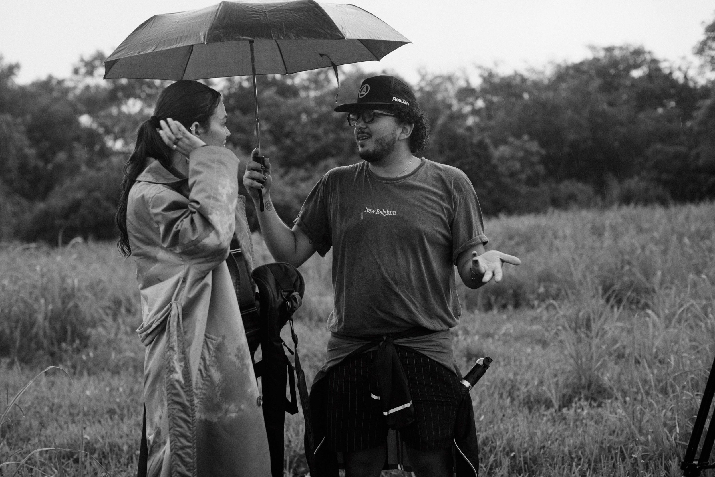 Two people talking outdoors in a field, one holding an umbrella. It is a black and white photo.
