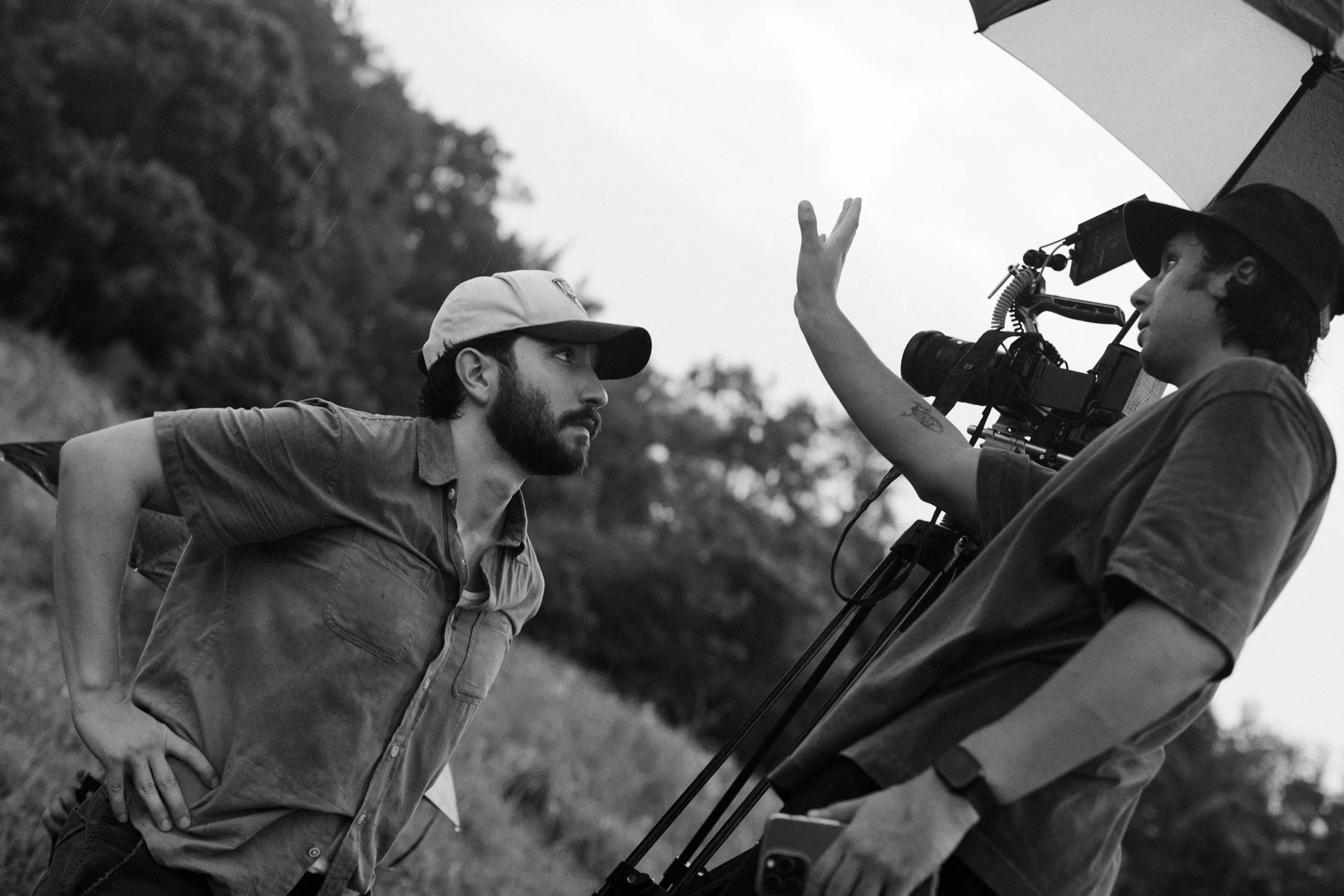 Two film crew members working outdoors, one wearing a baseball cap and the other a bucket hat, with camera equipment, an umbrella, and trees in the background.
