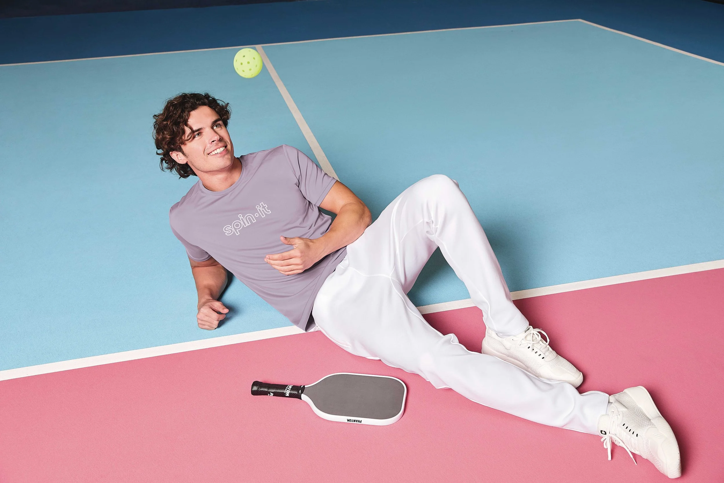 A young man laying on a colorful tennis court with a pickleball paddle and ball, wearing a lavender t-shirt with 'spin.it' printed on it and white pants.