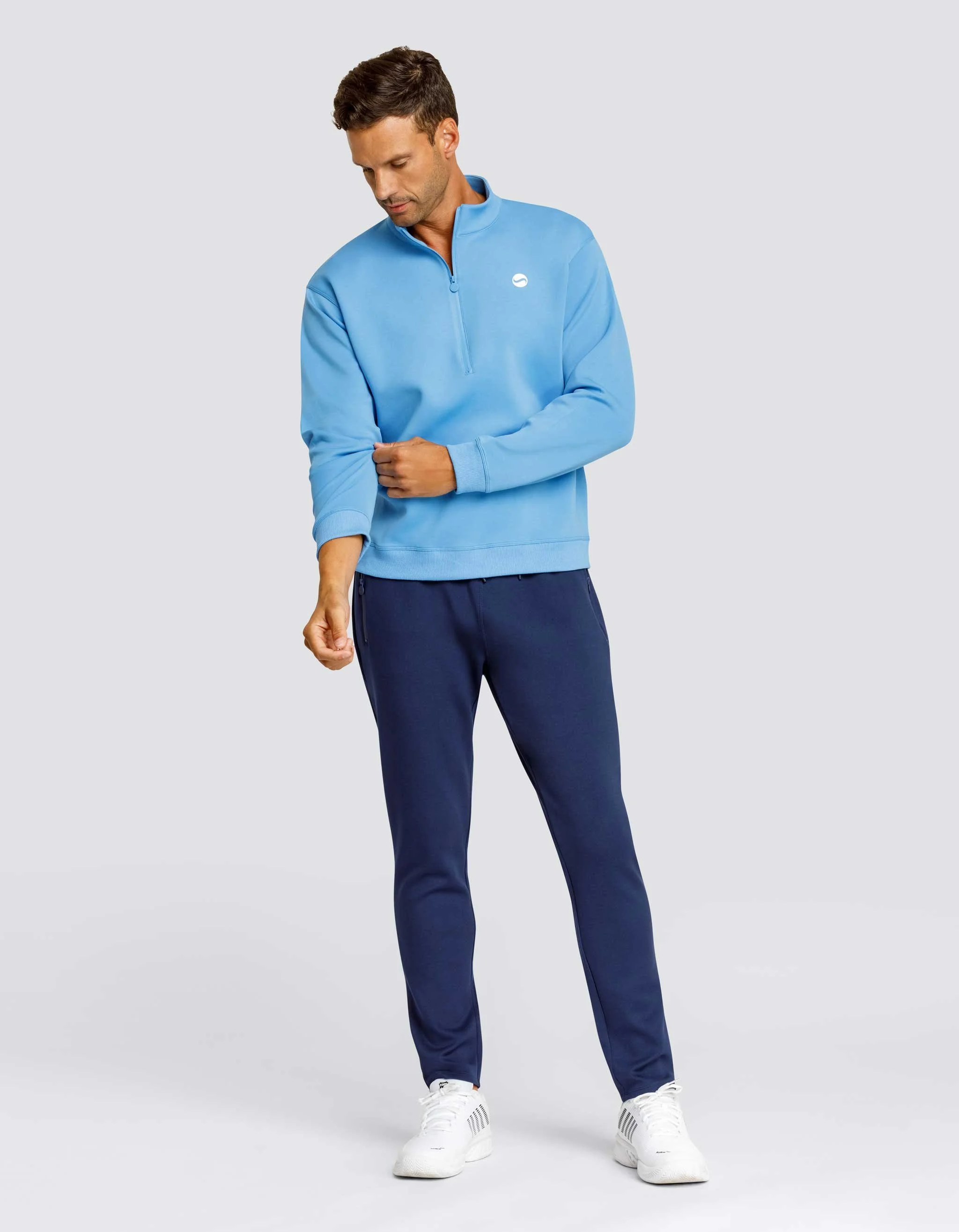 A man dressed in a light blue quarter-zip athletic top, navy blue track pants, and white sneakers standing on a plain gray background, looking down and adjusting his sleeve.