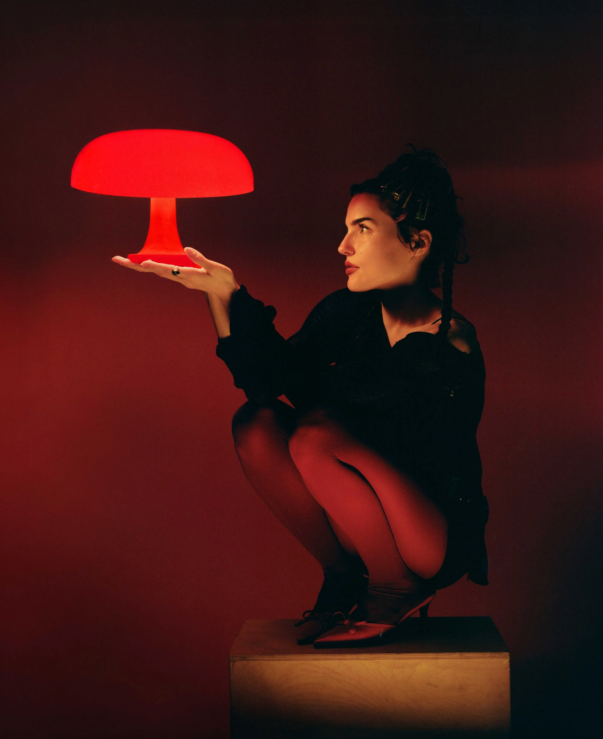 A woman with dark hair styled in bantu knots is sitting on a wooden block, holding a glowing red lamp in her hand. The background is dark, emphasizing the red glow of the lamp.