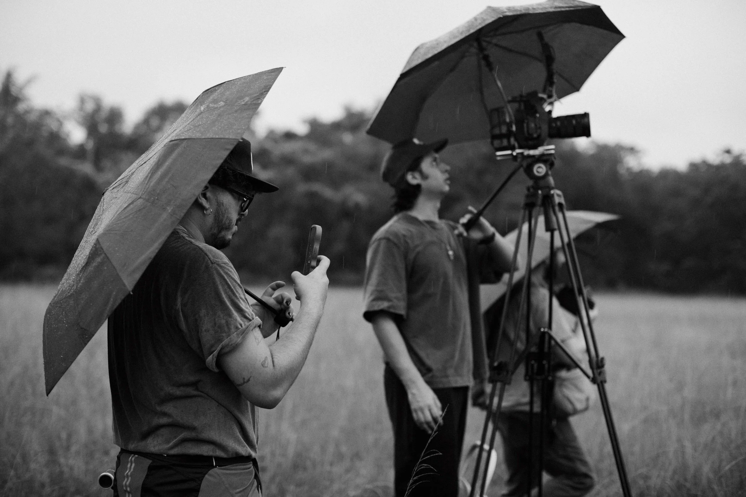 Three people with umbrellas and a camera on a tripod outdoors in overcast weather, possibly filming or photography.