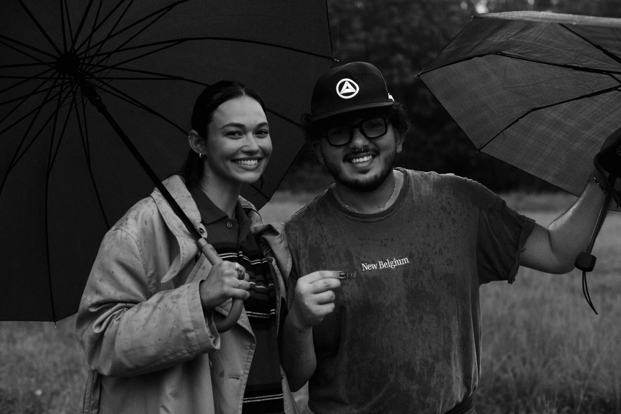 Two people smiling under umbrellas in the rain, one holding a drink and the other with a backpack, in an outdoor setting.