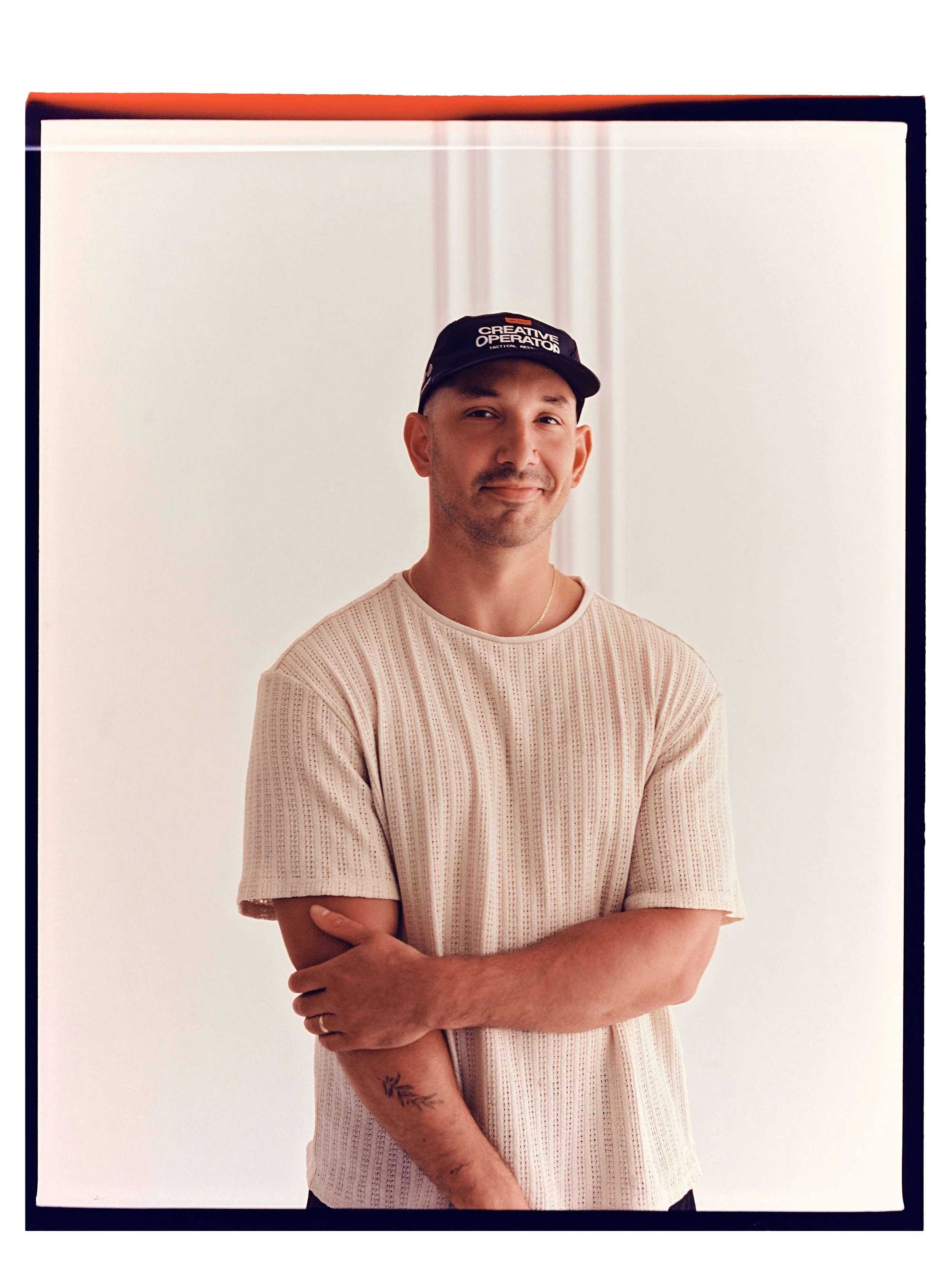 A man with a tattoo on his forearm, wearing a beige knit shirt and a black baseball cap with the words 'Creative Operator' written on it, standing against a plain white background.