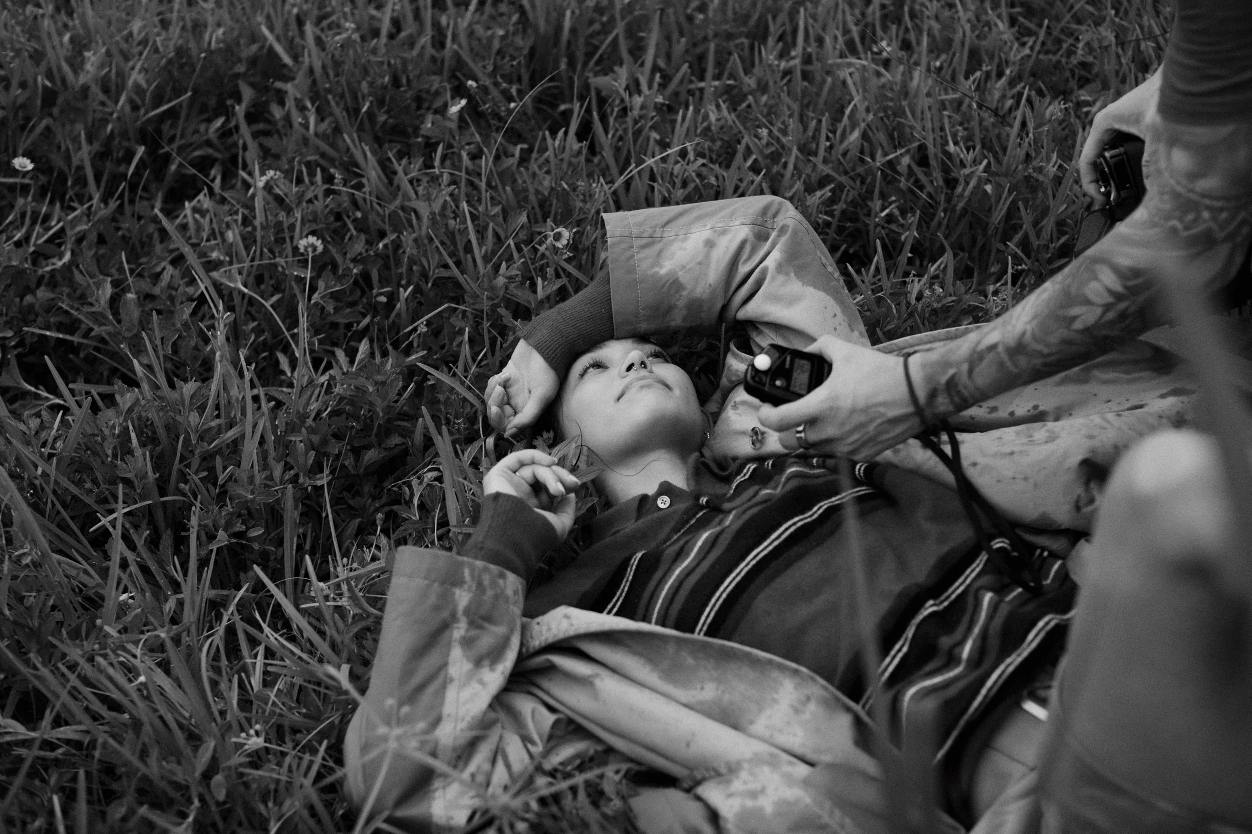 A person lying on grass with a child resting on their arm. The person is holding a camera, and the child is looking up, surrounded by small flowers in the grass.