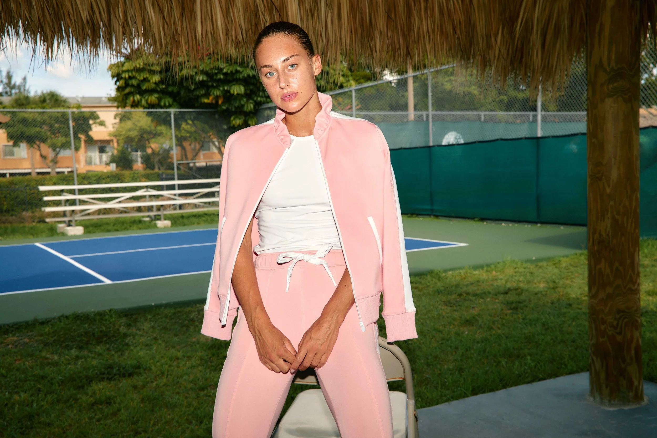 A young woman with blue eyes and brown hair slicked back, wearing a light pink tracksuit with white accents, standing outdoors near a tennis court under a thatched roof.