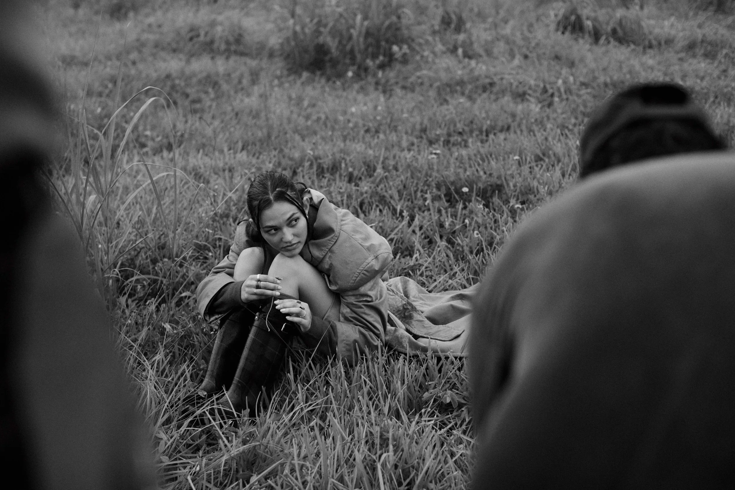 A black and white photo of a young woman lying on the grass outdoors, looking to the side with a serious expression. The photo is taken from a low angle, with blurred objects framing the image.