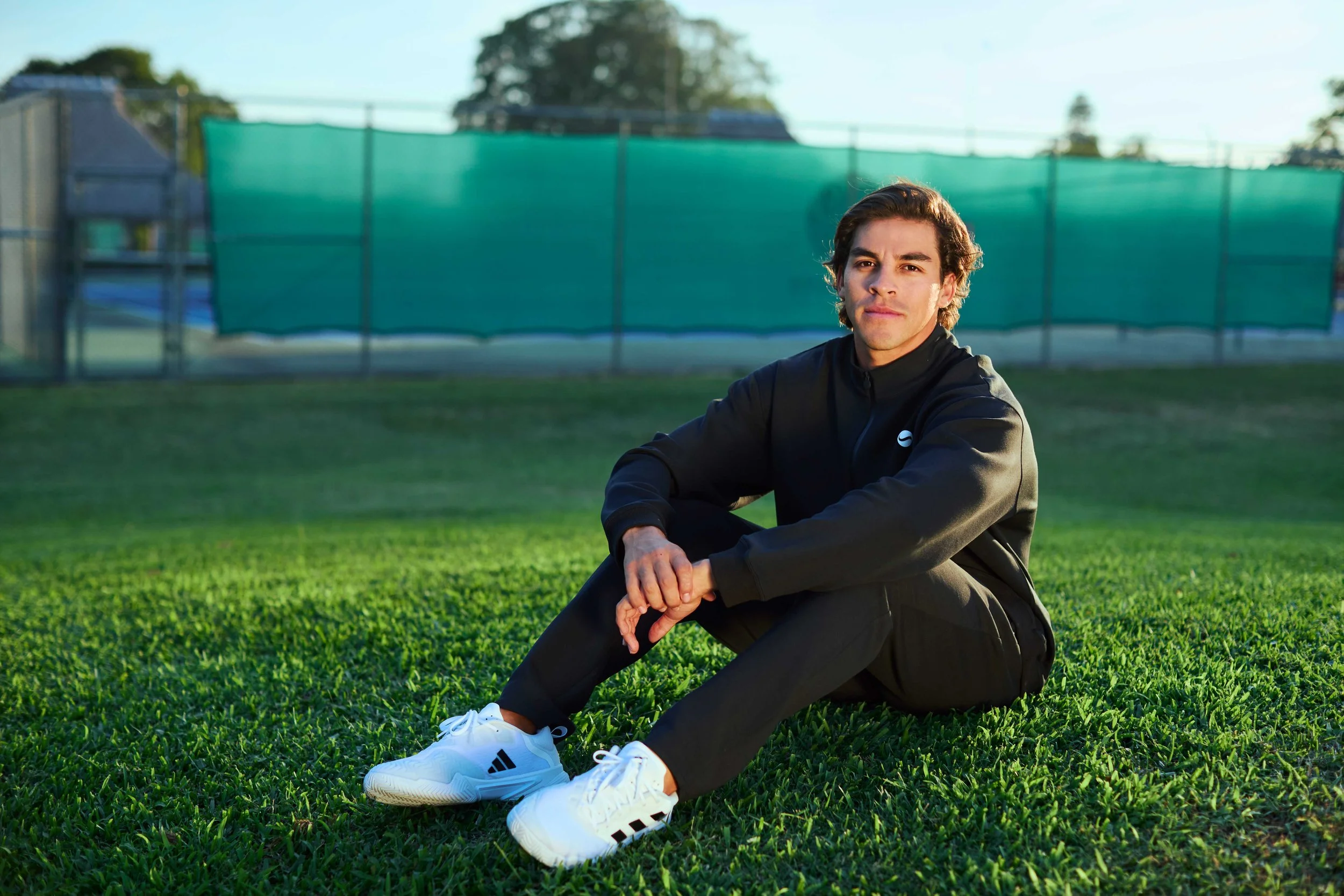 A young man with dark hair and tan skin is sitting outdoors on green grass, wearing a black athletic jacket, black pants, and white Adidas sneakers. He is looking at the camera with a neutral expression. In the background, there is a chain-link fence