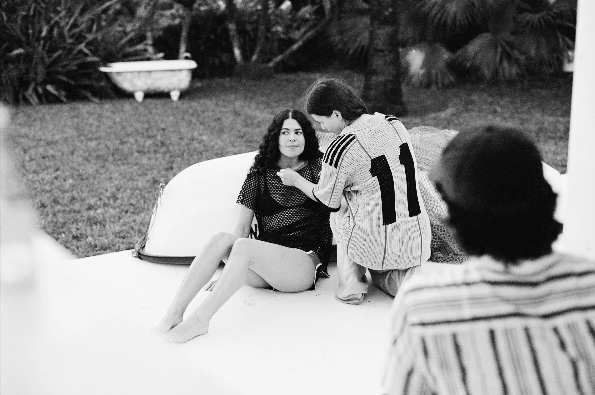 A woman seated outdoors on a white surface, wearing a patterned dress, and another woman leaning over her, adjusting her clothing or accessories, with a garden and plants in the background.