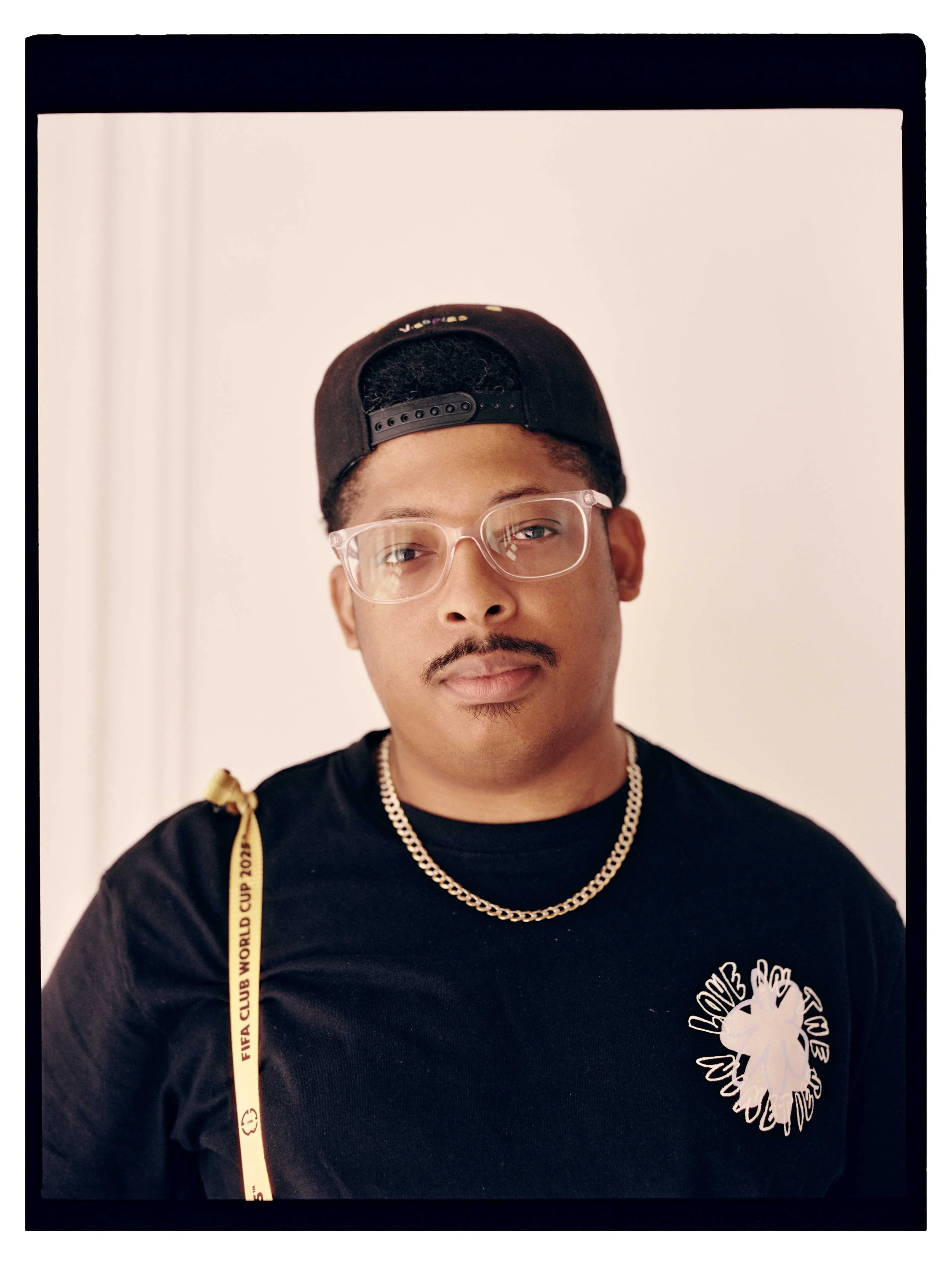 A young man with light brown skin, wearing a black baseball cap backwards, clear glasses, a black shirt with a white graphic, a gold chain, and a yellow lanyard with black text, standing against a plain, light background.