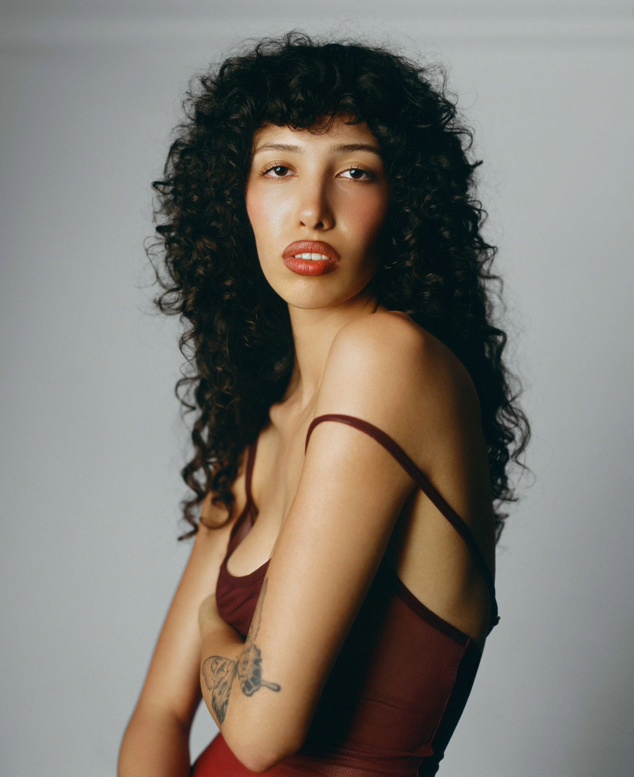 Portrait of a young woman with curly dark hair, wearing a dark red tank top, looking at the camera against a plain background.