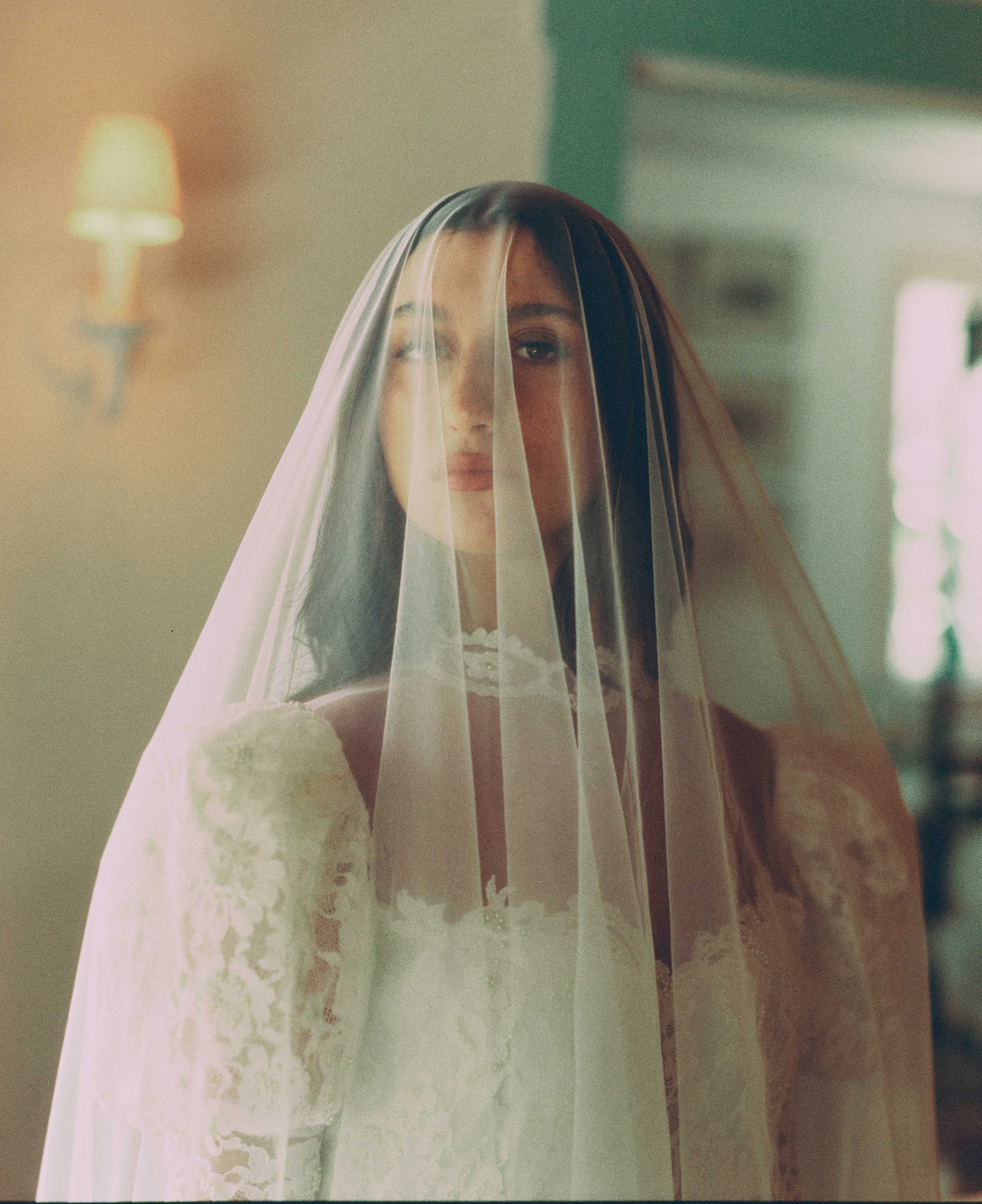 A woman with dark hair and a veil over her face, standing indoors with a soft-focus background and warm lighting.