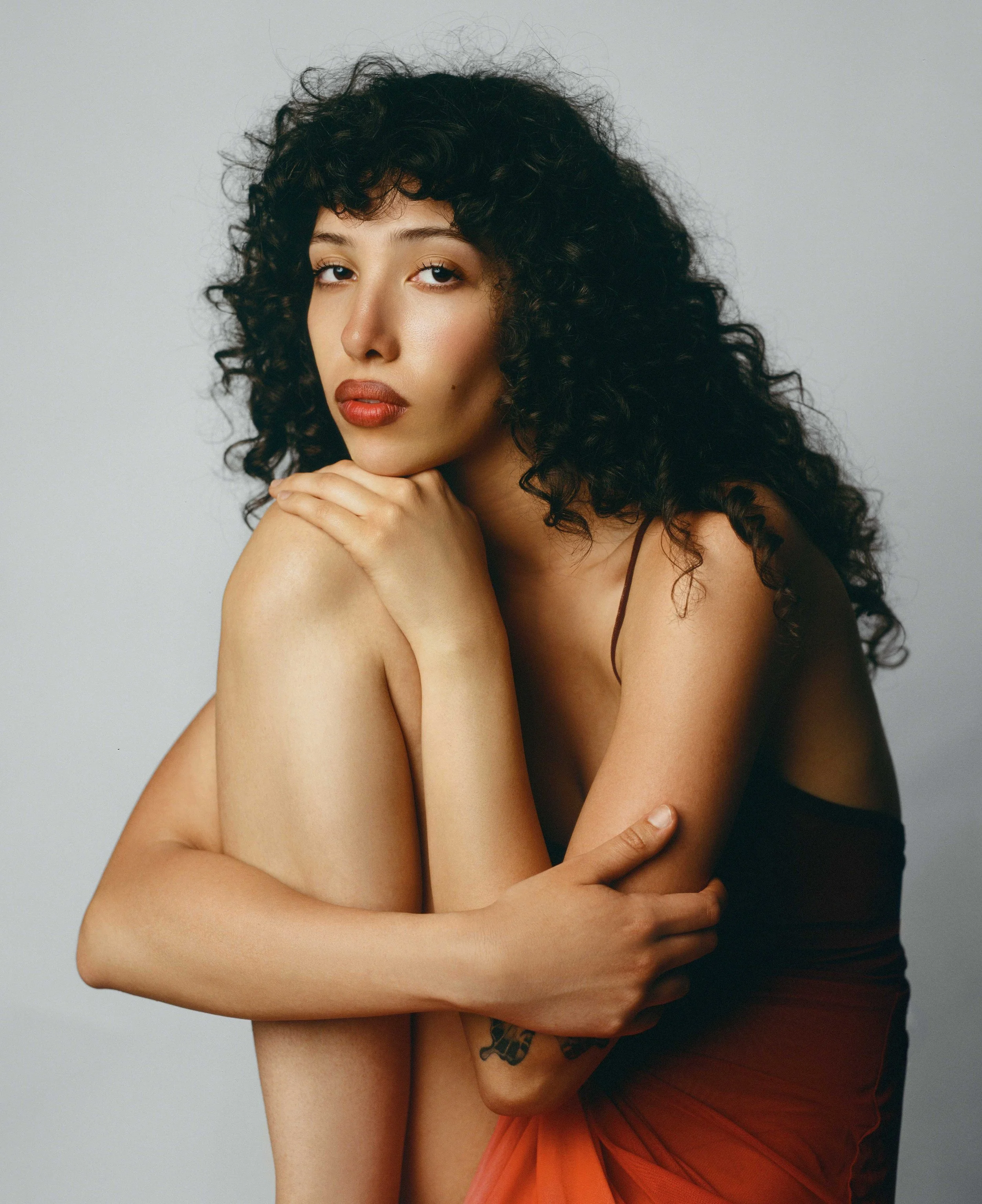 A woman with curly dark hair and natural makeup poses with her arms crossed and resting on her shoulder, wearing a sleeveless top and a sheer orange skirt.