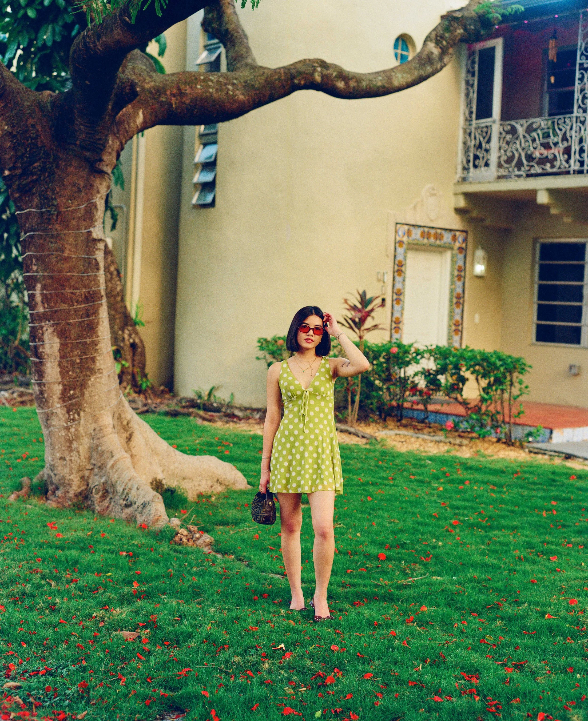 A woman with black hair, wearing red sunglasses, a light green polka-dot dress, and holding a designer handbag, walking on a green lawn with red flowers in front of a yellow building with balconies and plants.