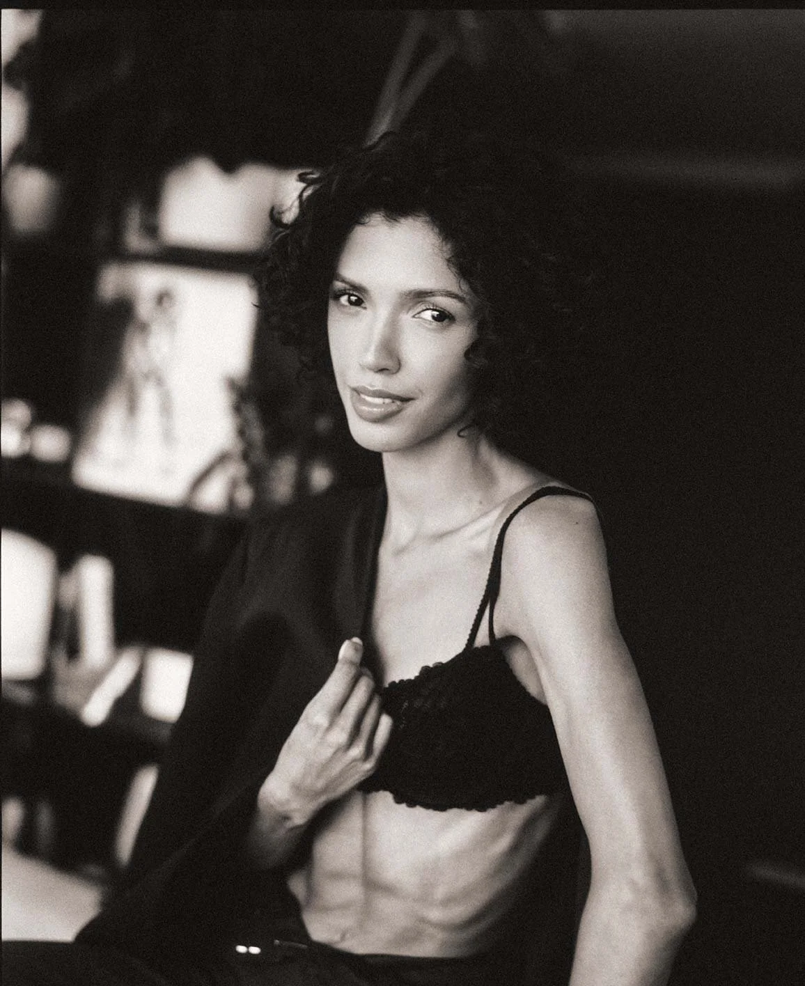Black and white photo of a woman with curly hair, wearing a bra and a jacket draped over her shoulder, posing indoors with a slight smile.