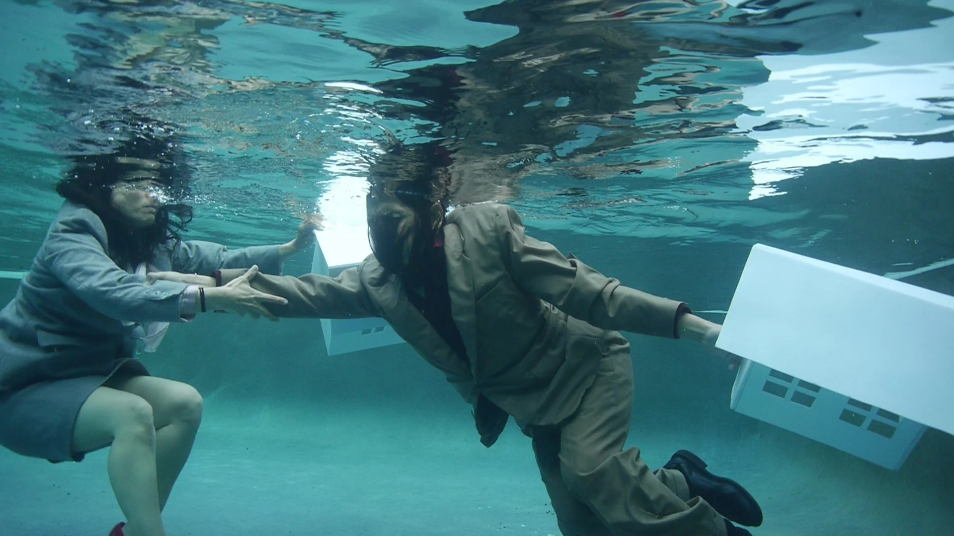 Two people wearing suits underwater reachout a hand to each other