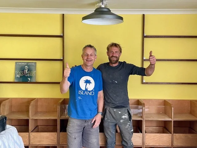 Two men standing in front of wooden shelves against a yellow wall with a poster of Frankie Laine's "Rawhide" song."