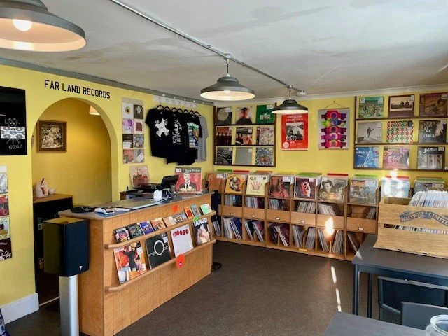 Interior of a small record store with yellow walls, featuring vinyl records displayed on shelves, a wooden checkout counter with various records, T-shirts hanging on the wall, and posters framed on the walls.