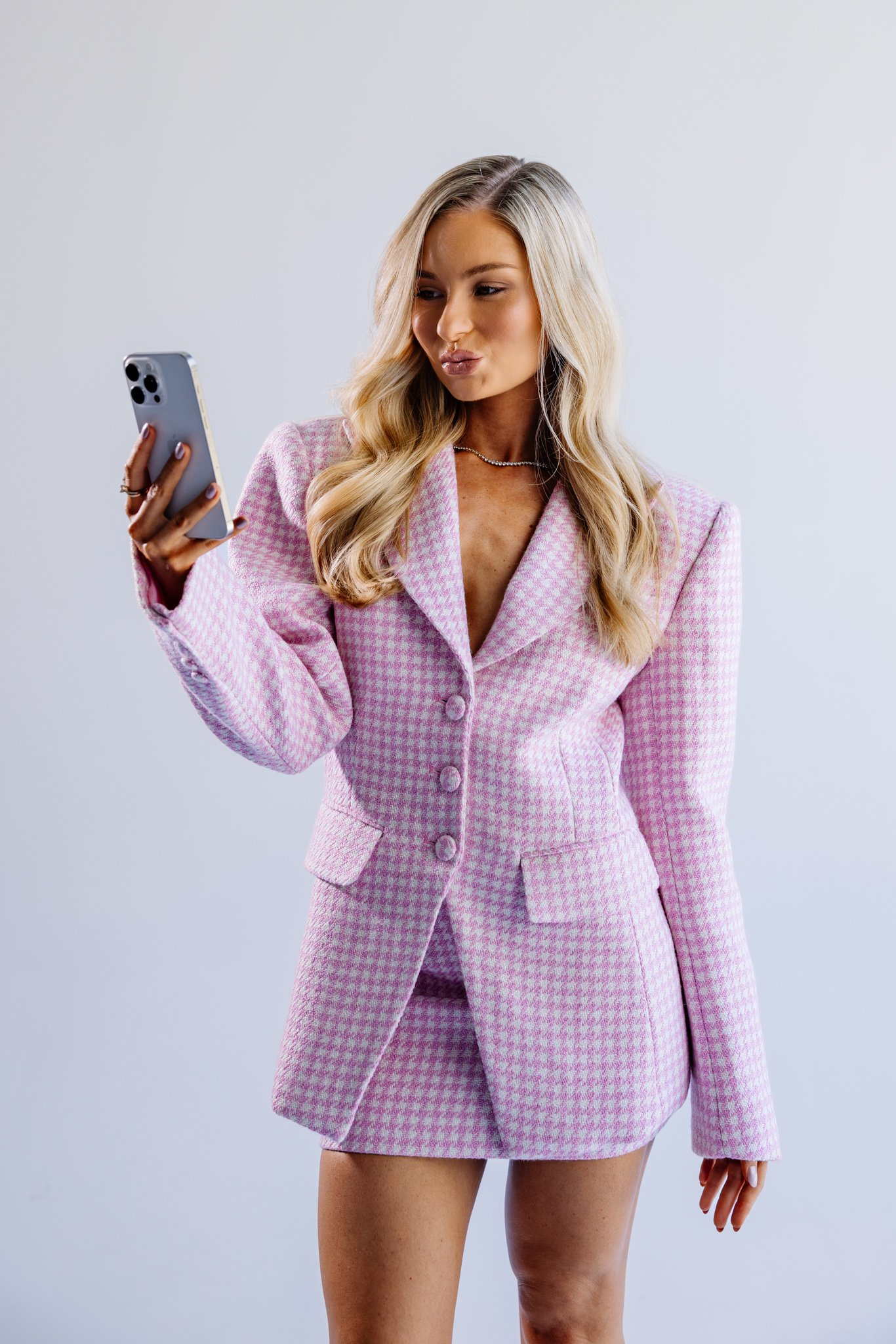 A woman with blonde hair in loose waves wearing a pink checked blazer and matching skirt, looking at her phone and pursing her lips.