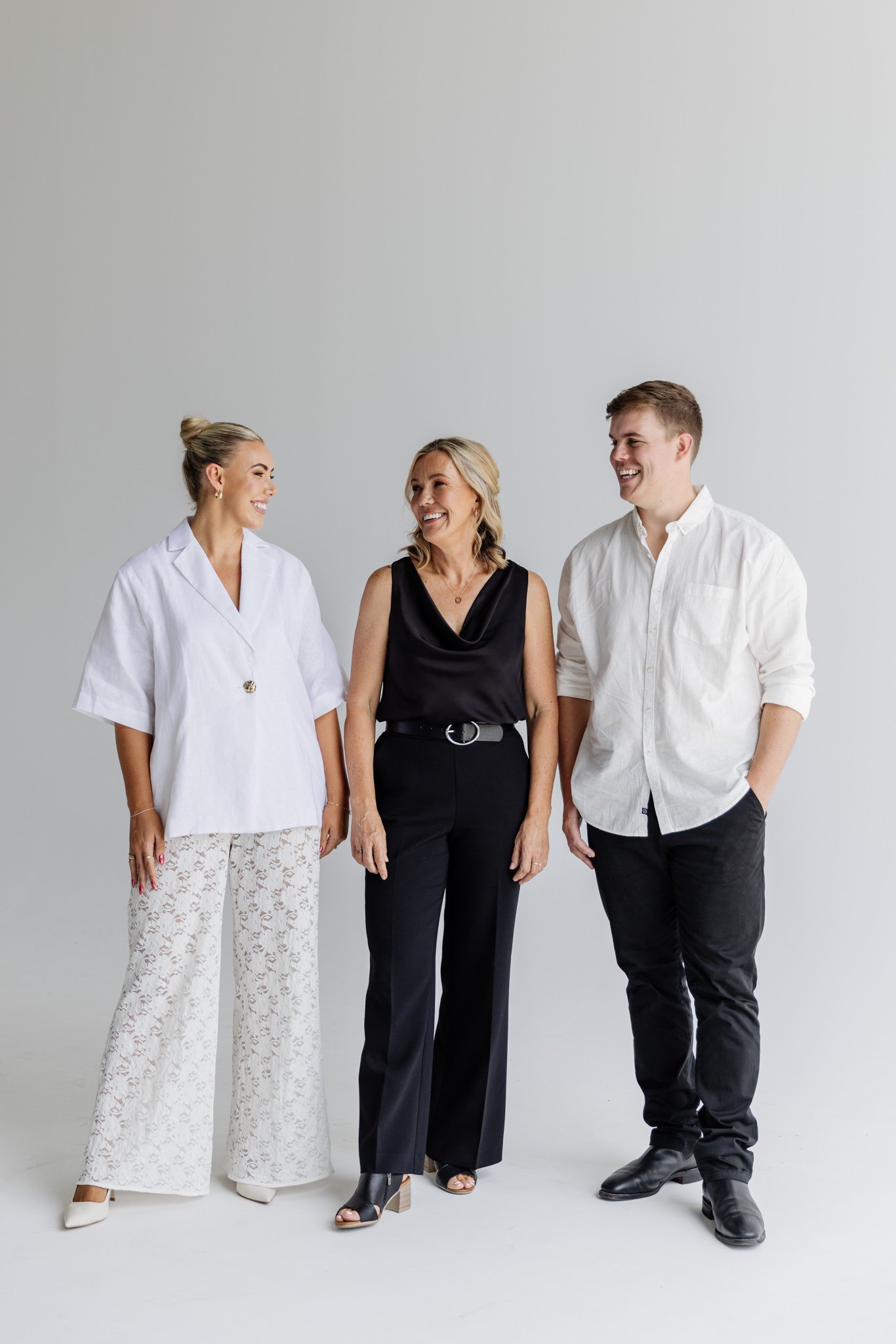 Three people standing and smiling in front of a plain light gray background, dressed in business casual attire