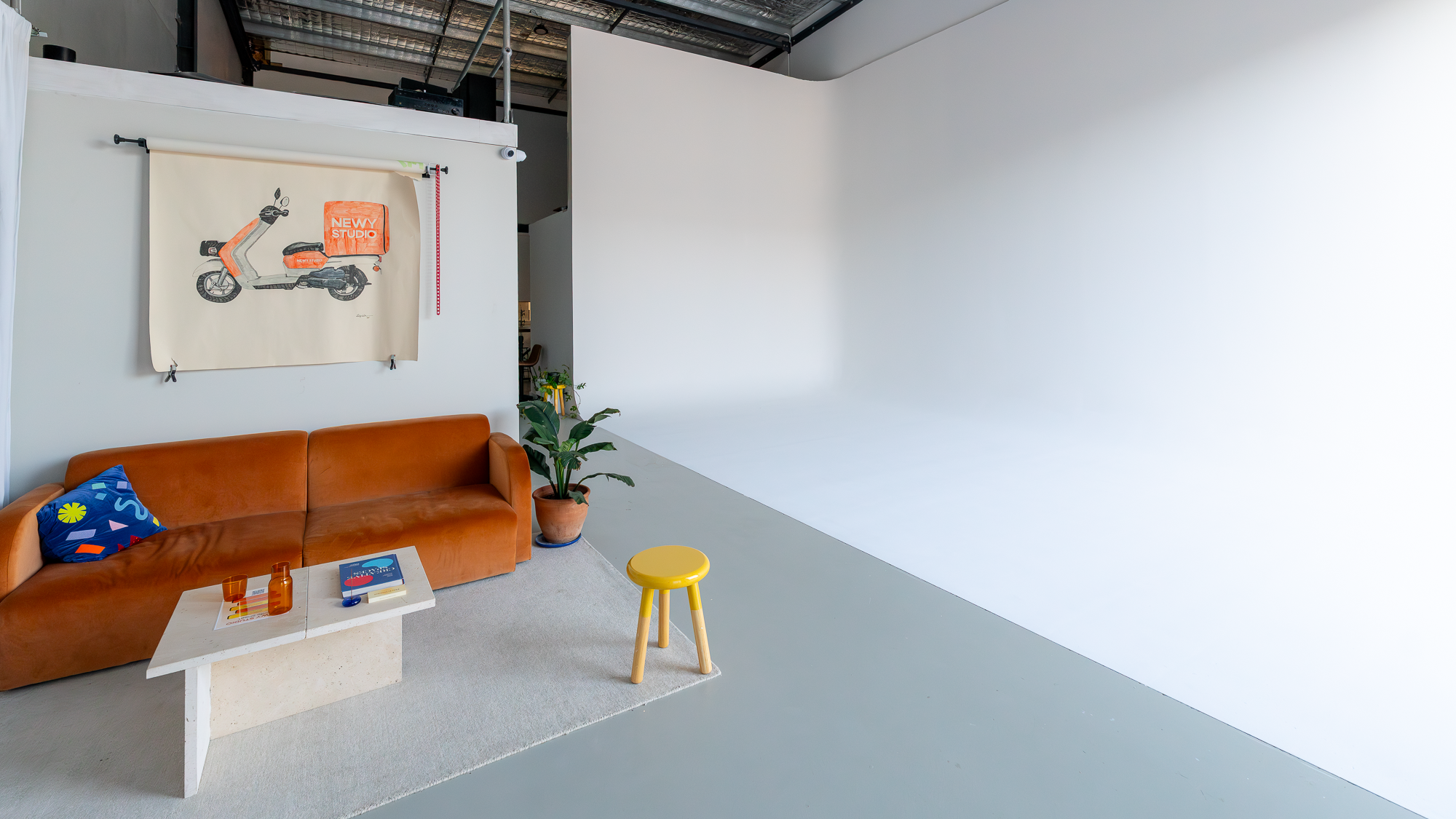 A modern studio space with a tan leather sofa, a small white coffee table with orange glasses and a book, a yellow stool, a large potted plant, a decorative curtain with a scooter illustration, and a plain white wall.