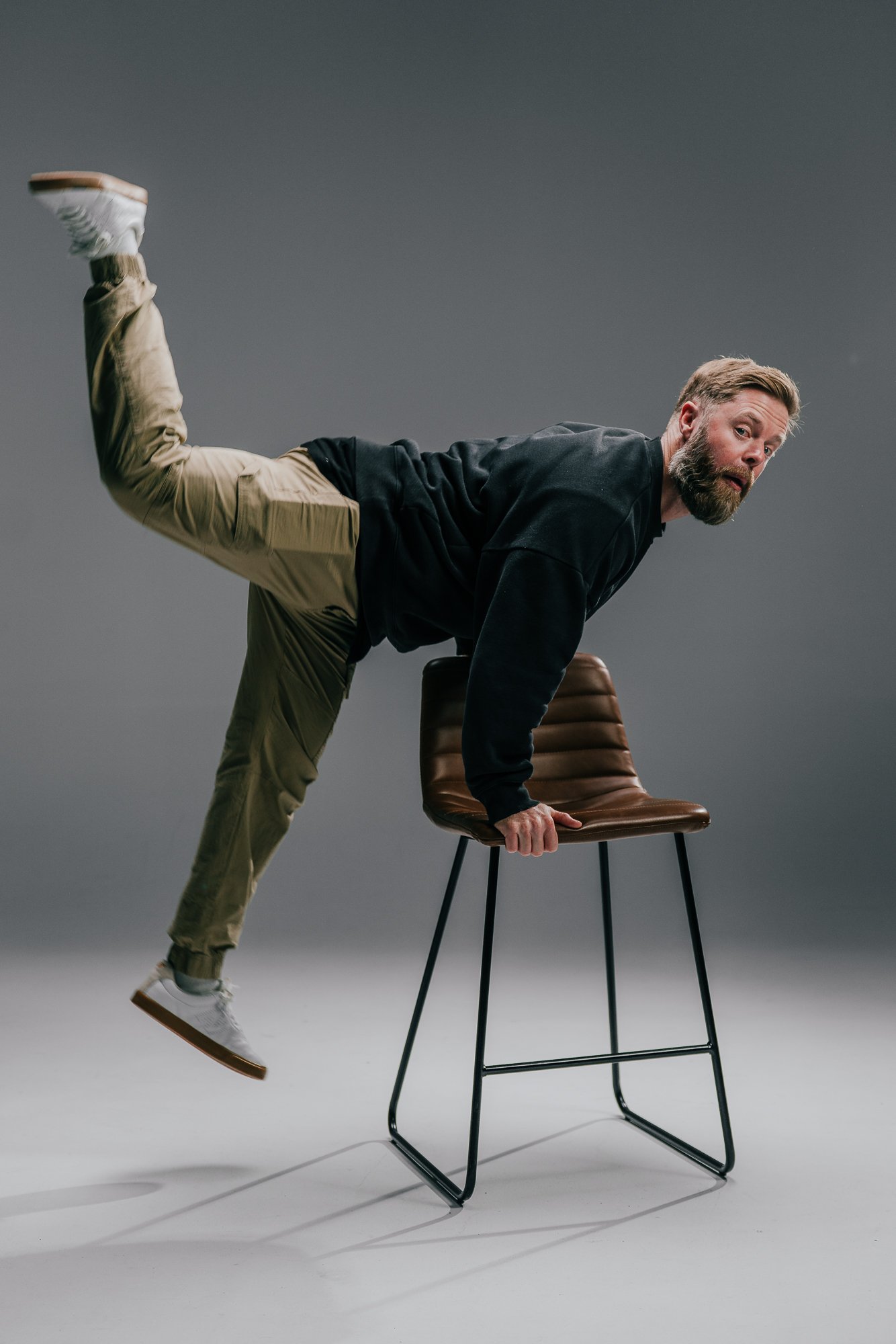 A man balancing with his hands on a chair, one leg extended behind him, against a plain gray background.