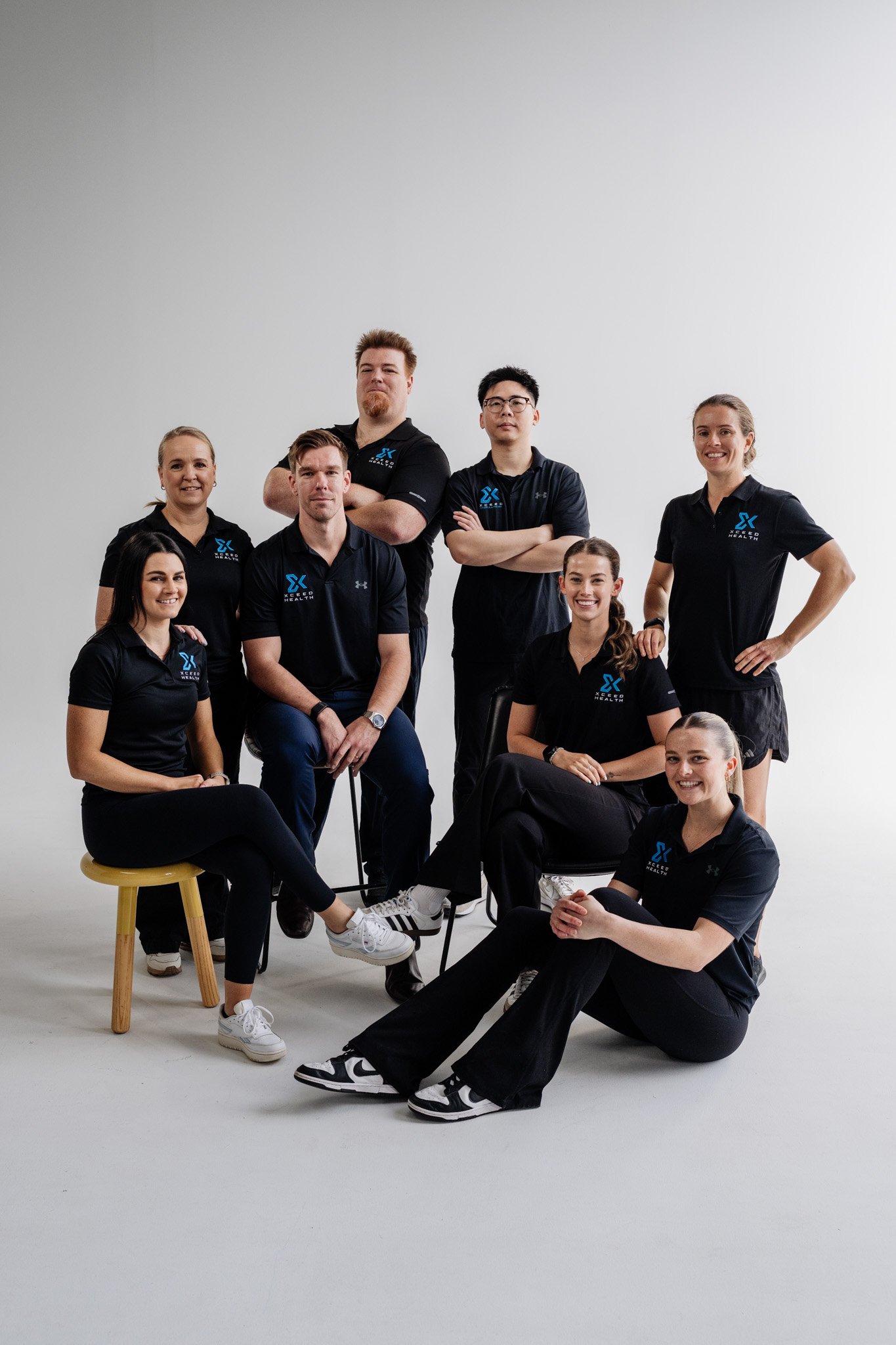 Group of eight people in black polo shirts with blue logos, posing in a studio with a plain white background.