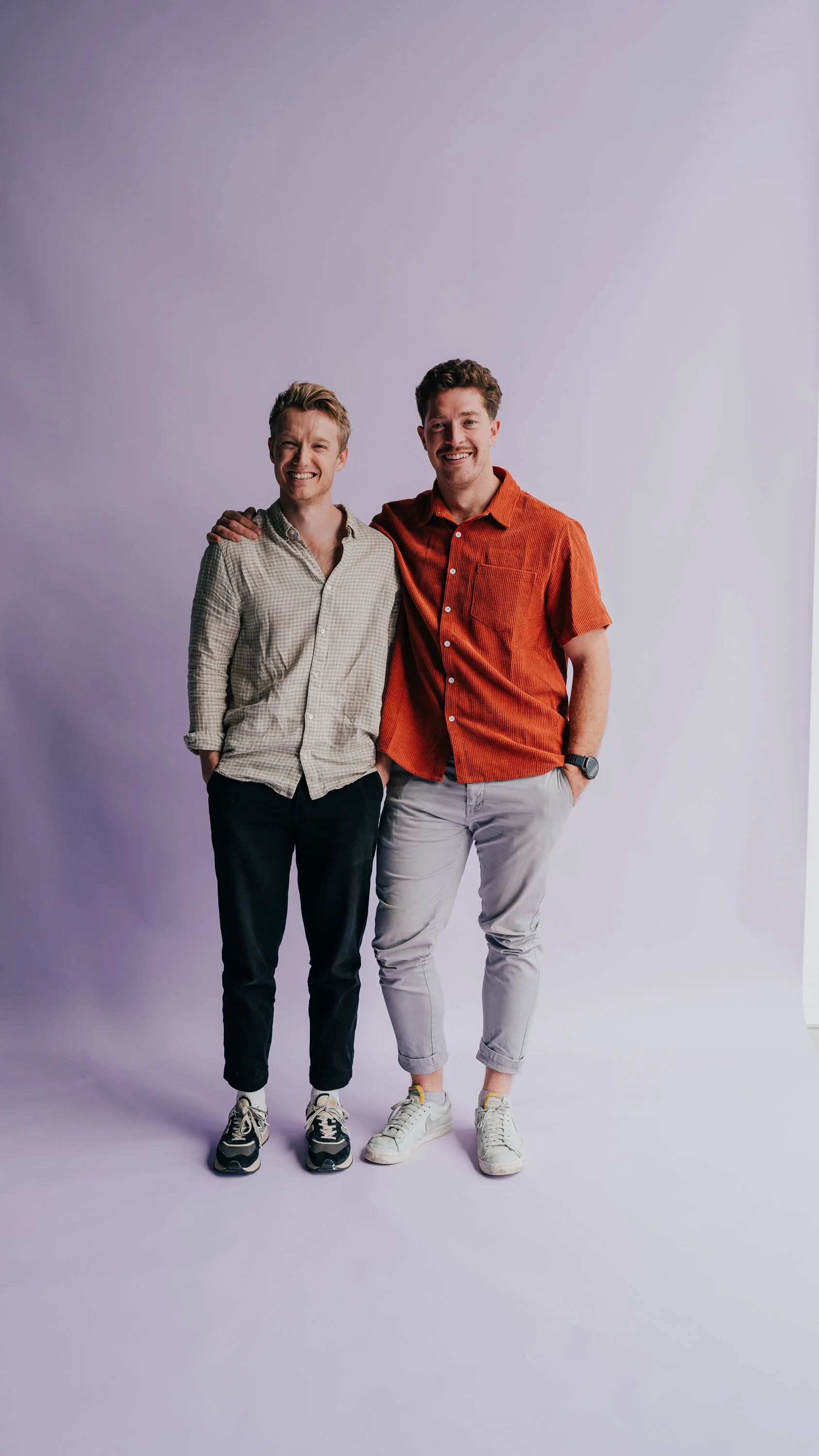Two men standing together on a light purple studio backdrop, smiling, with one arm around each other's shoulders.