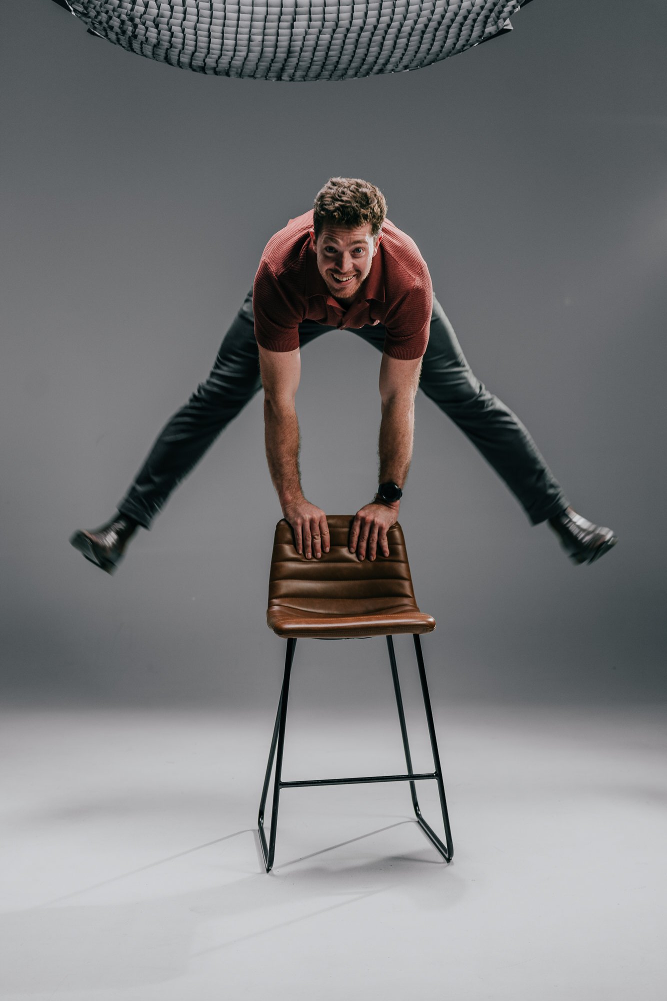 A man in a red shirt and black pants performing a handstand on a chair, with his legs spread apart, smiling, in front of a plain gray background.