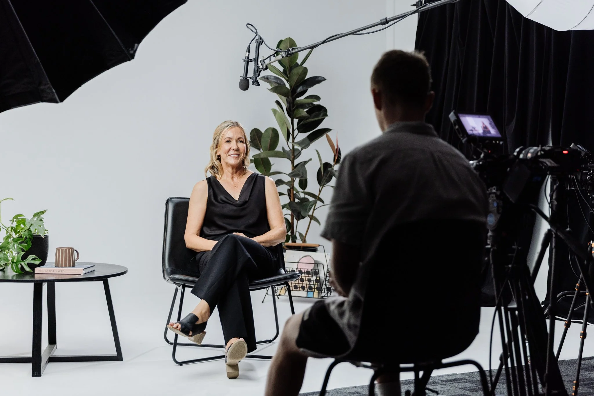 A woman in a black sleeveless top and black pants sitting on a black chair, being recorded by a man with a camera in a studio setting with a white background, black curtains, a large plant, and a small table with books and a plant.