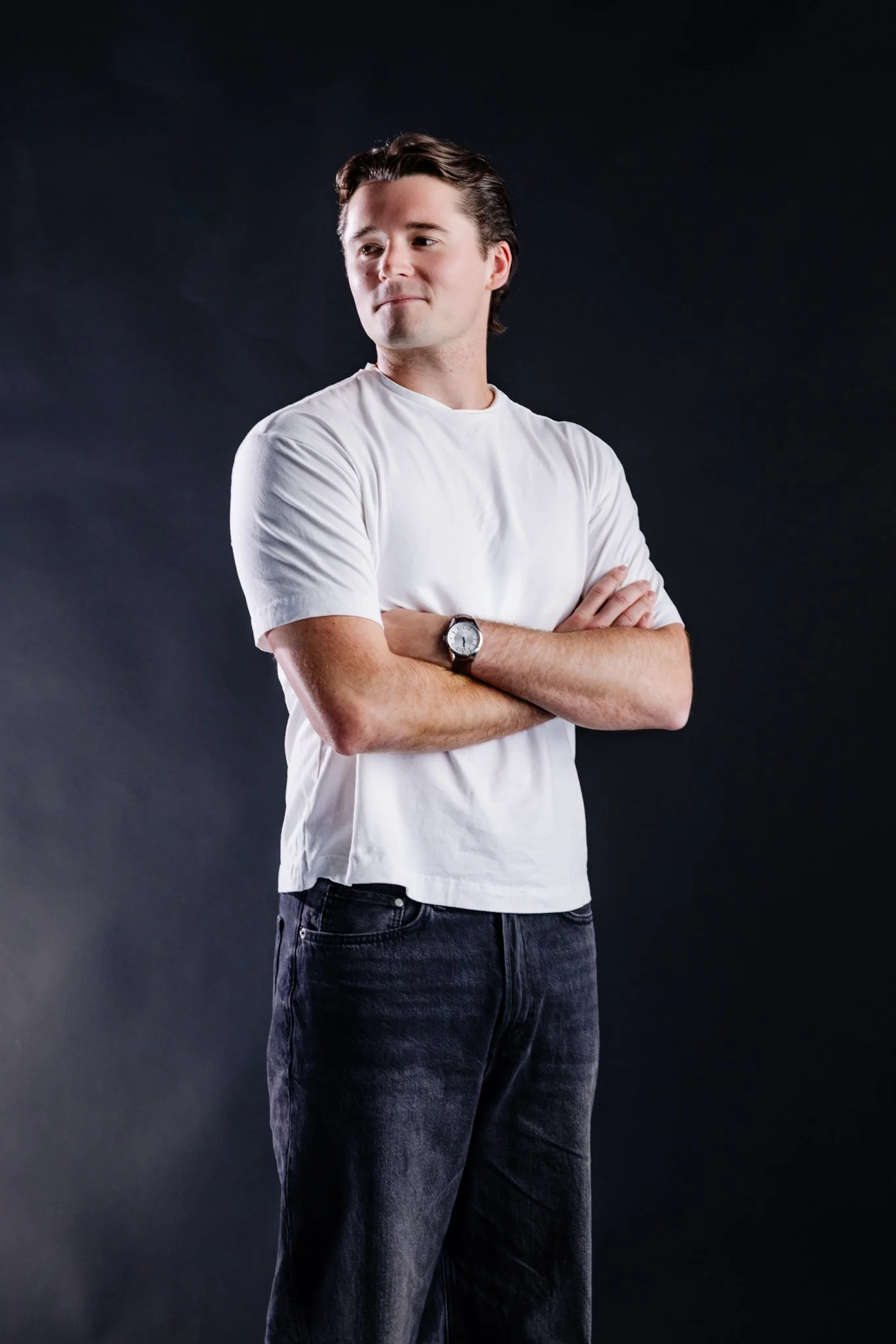 A young man with brown hair, wearing a white t-shirt and black jeans, stands with his arms crossed against a dark background.