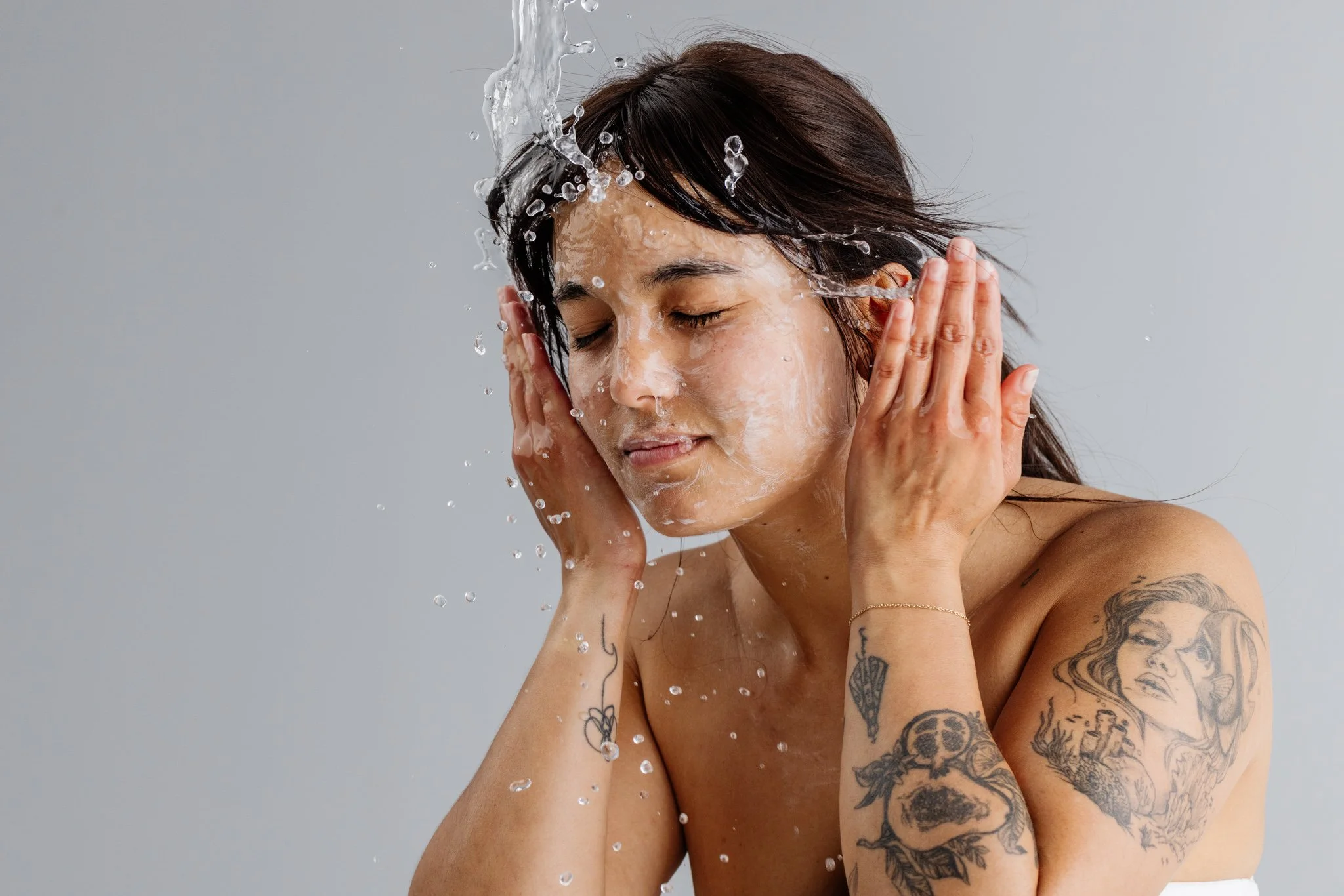 A woman with shoulder-length dark hair and tattoos on her arms, washing her face with water, with water splashing over her head and face.